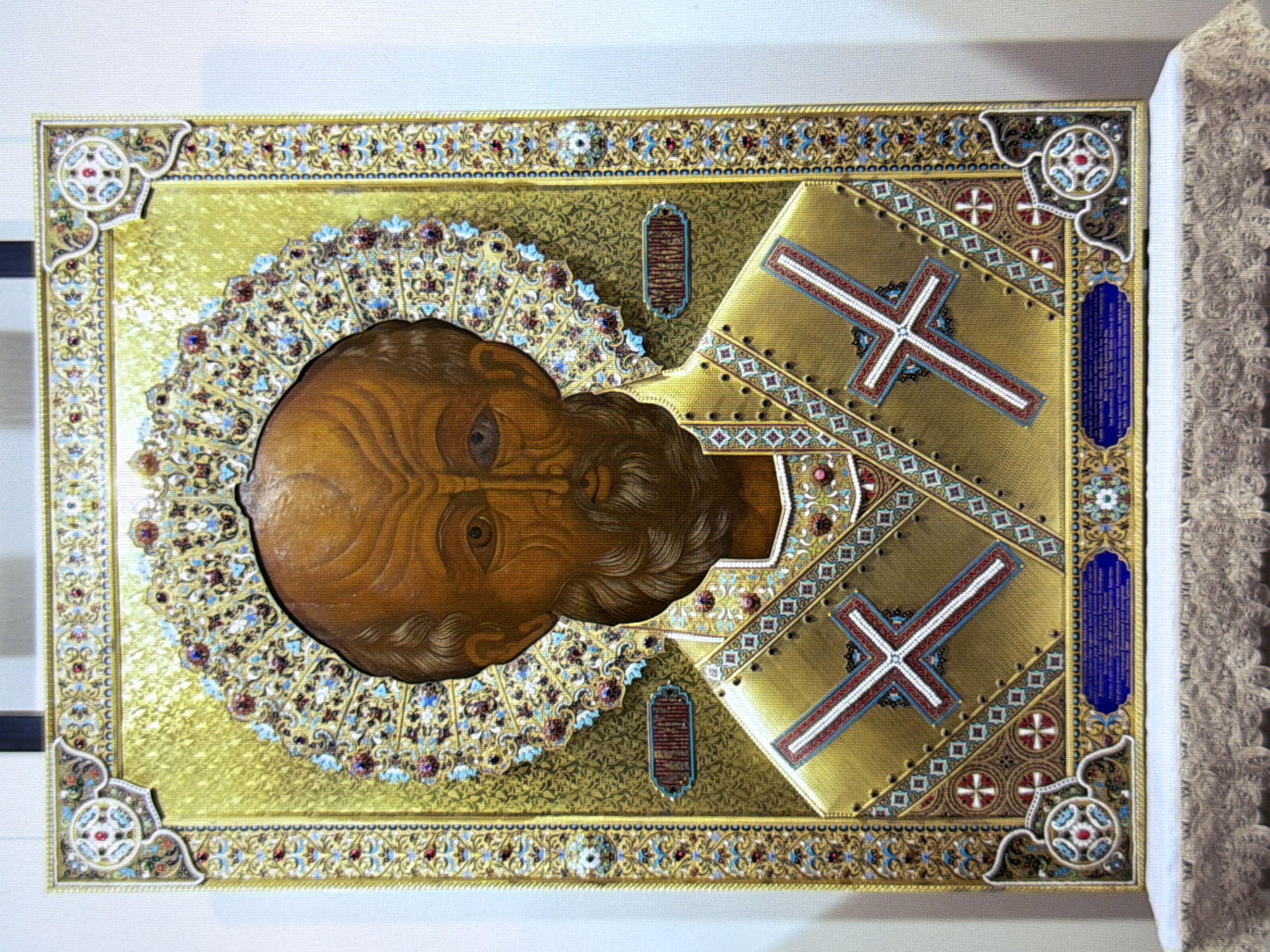 The image shows an icon of Saint Nicholas, depicting his bust and upper torso, framed by an ornate, jeweled halo and elaborate golden vestments. The background is a textured gold. Two prominent crosses are embroidered on his garments. The icon is presented as a physical object, likely for display or veneration, and is propped on a lace-covered surface. Text is visible on the icon in what appears to be Cyrillic script. The background environment suggests an indoor setting, possibly a museum or church.
The icon is located in the National Museum of the History of Ukraine in the city of Kyiv, Ukraine.