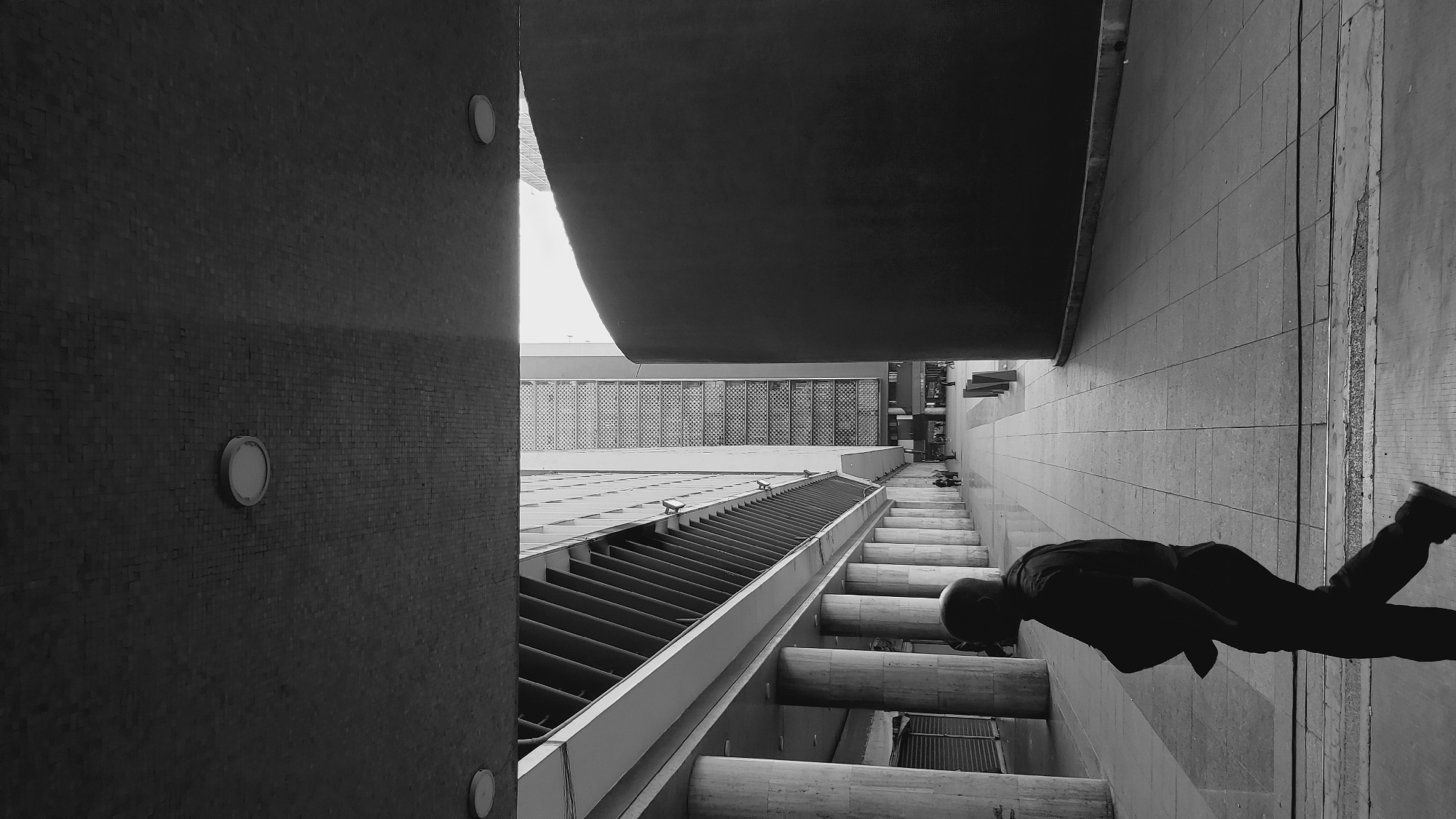 The black and white photograph shows an exterior architectural space in Caracas, Venezuela. A wall is located on the left and the edge of an overhanging, curved structure is situated at the top. The perspective is directed downwards into a narrow, open area between buildings. The buildings feature cylindrical pillars and horizontal slatted structures. A silhouette of a person is on the right, seemingly lying down on the ground.