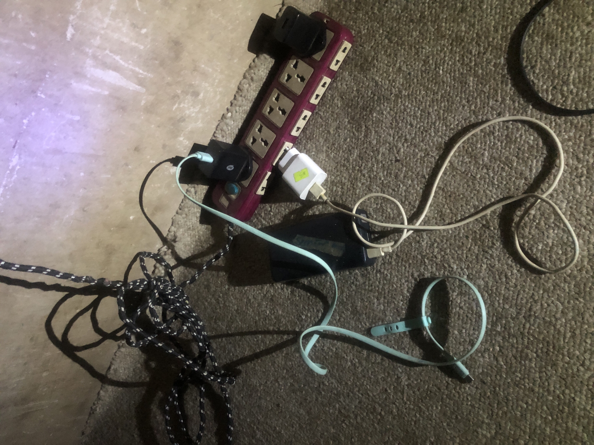 The image displays a power strip with multiple electrical plugs connected to it, along with various charging cables and devices scattered on a carpeted floor. A black and white braided cable is prominent, looping around some of the other items. There's a black electronic device, possibly a power bank or a tablet, with a light blue charging cable attached. Another charging cable, white in color, is also visible. A light blue wristband with visible holes is laid out on the floor. The power strip is a multi-socket universal adapter, purple in color, with several black and one white power adapter plugged into it. The background shows a textured wall with faint scratch marks and some illuminated areas, suggesting artificial lighting. The scene is located in Jalingo, Nigeria, and appears to be an indoor setting, likely a room. No people are present in the image, and no explicit actions or interactions are depicted beyond the state of connected and unconnected electronics. The overall impression is of a cluttered charging station.