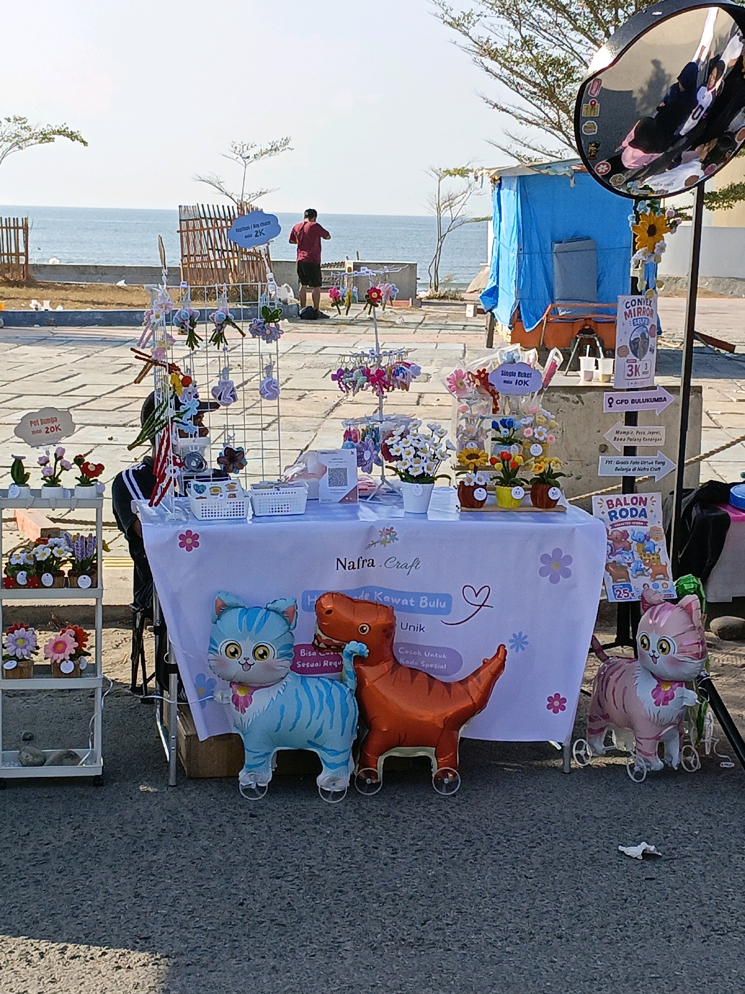 This image depicts an outdoor stall selling crafts and balloons in Bulukumba, Indonesia. The stall is set up near the coast, with the ocean visible in the background. A man in a red shirt and dark shorts is standing near the water, facing away from the camera.
The stall itself is decorated with various items, including flower crafts, balloons in the shapes of a blue cat, an orange dinosaur, and a pink cat. A sign on the table reads "Nafra Craft" and also features text in Indonesian that translates to "Soft Fur Cat," "Unique," and "Suitable for Special Gifts." Another sign behind the stall advertises "BALON RODA" (Wheel Balloon) for 25,000 IDR. There are also various price tags visible, with some items priced at 2K, 10K, and 20K.
The environment suggests a sunny day with a clear sky. The stall is located on a paved area, likely a boardwalk or promenade, adjacent to a sandy area and the sea. The overall scene captures a commercial setting with a relaxed, possibly tourist-oriented atmosphere.