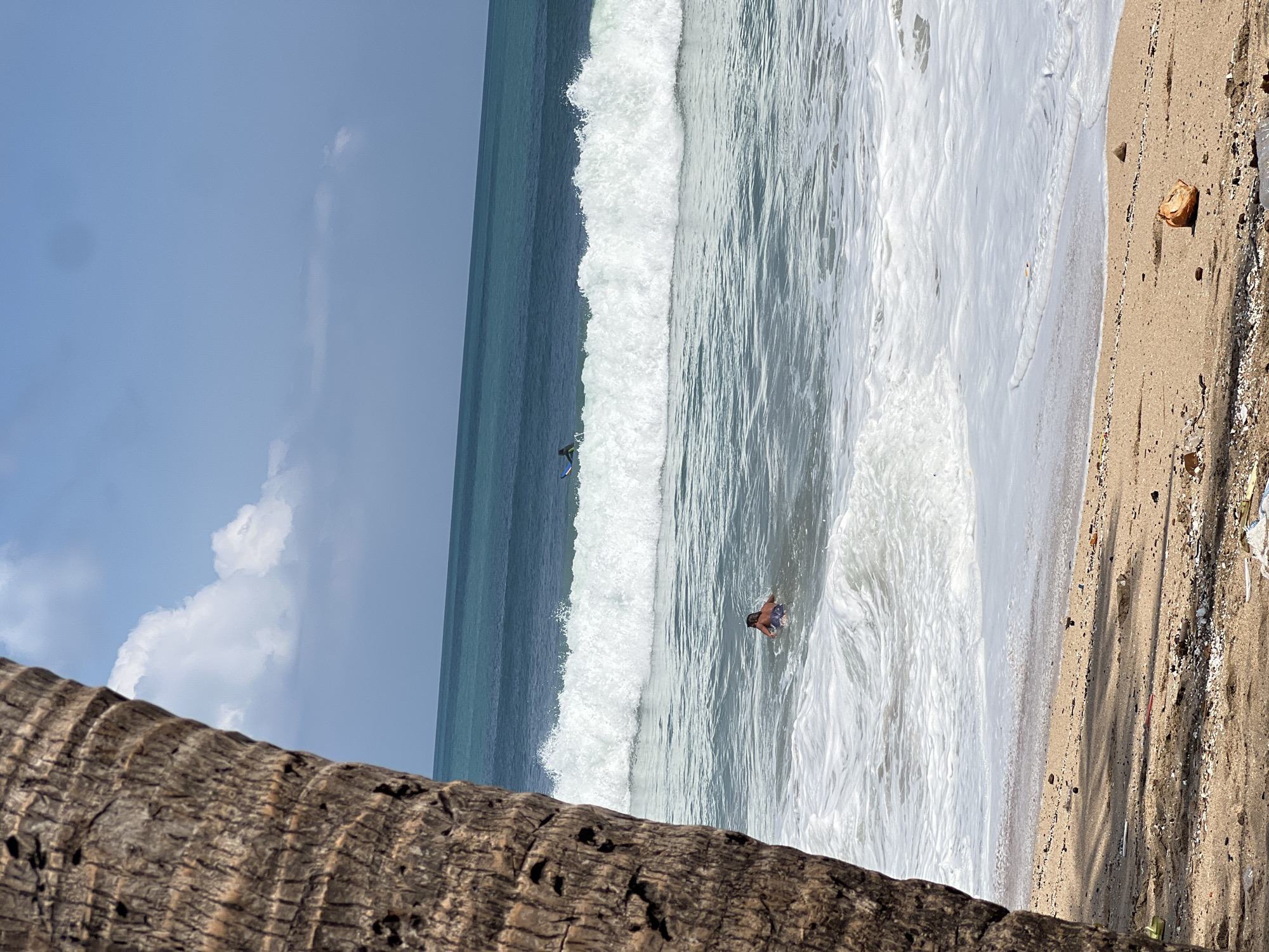 A vertical, close-up shot of a palm tree trunk in the foreground of a beach scene in Kuta, Indonesia. The trunk is rough and textured, with dark brown bark. In the background, the turquoise ocean stretches to the horizon under a bright blue sky with scattered white clouds. Two surfers are visible in the water; one closer to the shore is swimming, while the other is further out, riding a wave. The beach in the foreground is sandy and dotted with small shells and debris.