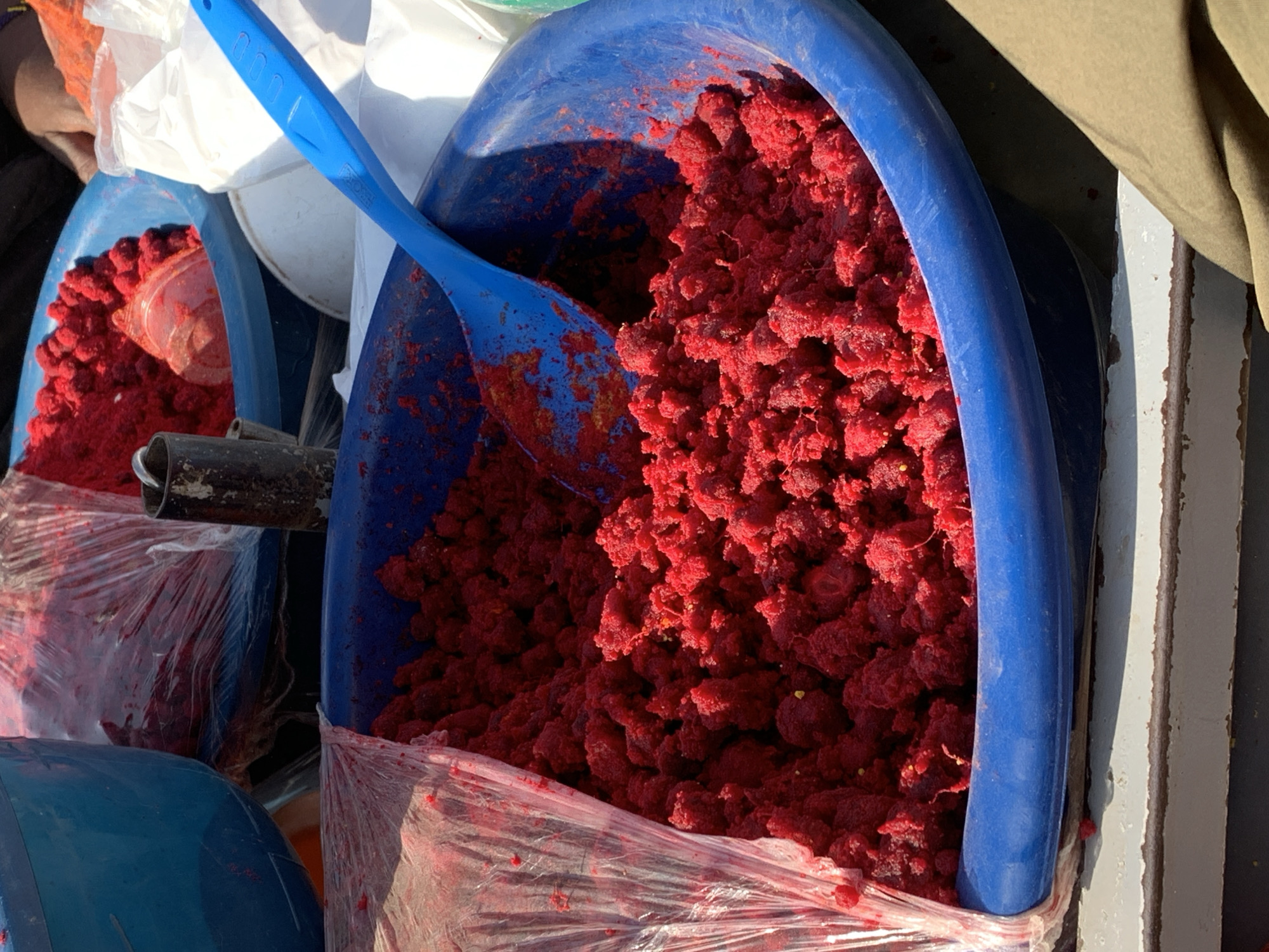 The image shows a close-up of a blue plastic bowl filled with a bright red, granular substance, possibly crushed berries or a spice. A blue plastic spoon is resting inside the bowl, with some of the red material clinging to it. The bowl is partially covered with clear plastic sheeting. In the background, other blue containers, a white bag, and a glimpse of a person's hand are visible, suggesting a market or food preparation setting. The scene appears to be outdoors under natural light, casting shadows. The foreground shows a weathered surface, likely a table or counter. The overall impression is of a vibrant, colorful, and somewhat rustic display of ingredients.