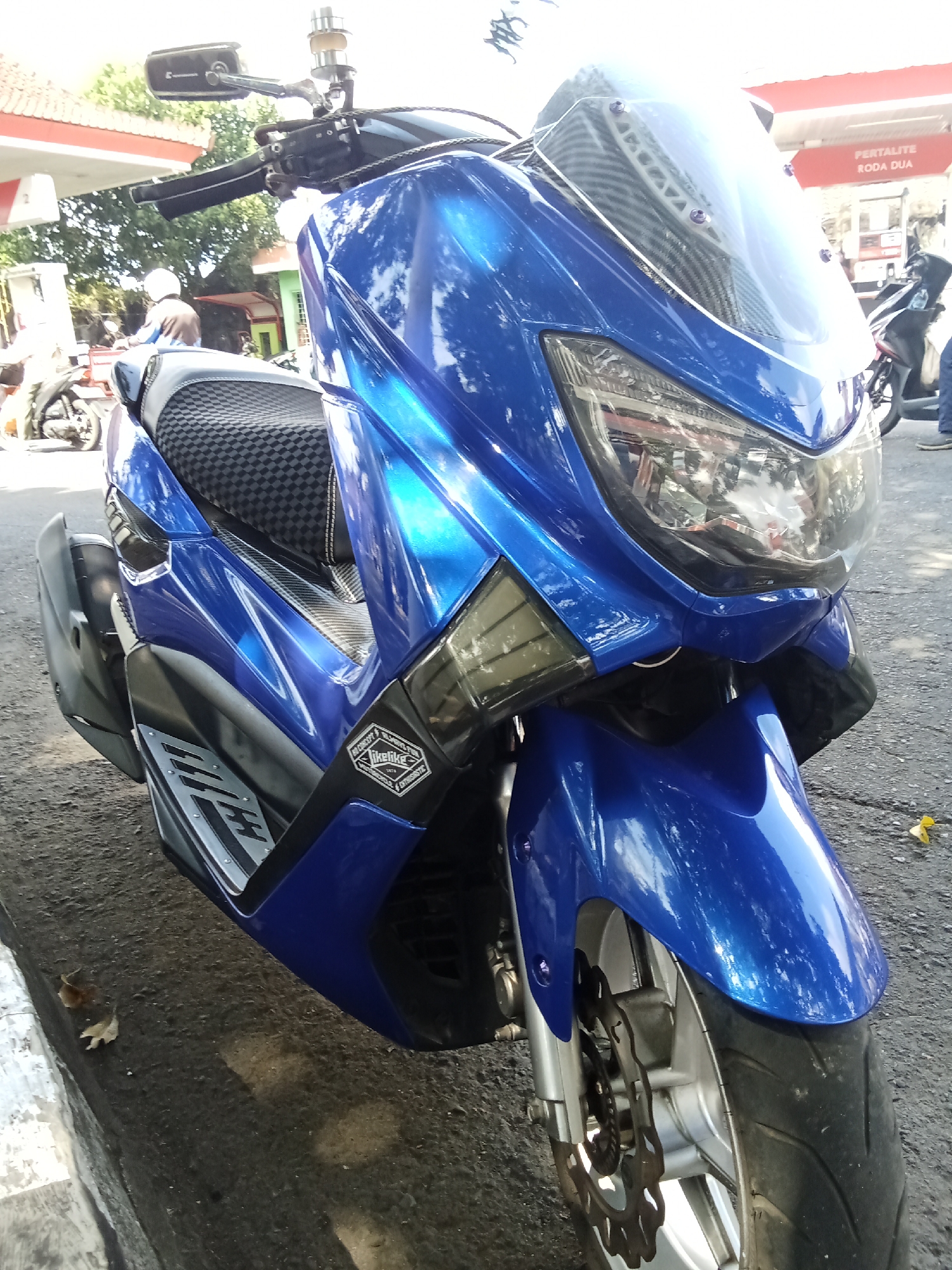 The image shows a blue motorcycle parked on an asphalt surface, likely a street or parking lot. The motorcycle is the primary subject, with its front wheel, handlebars, seat, and part of the fairing visible. The motorcycle has a reflective blue finish, a black seat with a checkered pattern, and a clear windscreen.
In the background, there is a building with a red and white awning, which appears to be a gas station. A sign on the awning reads "PERTALITE RODA DUA". There are also some trees and other motorcycles visible in the background, suggesting an urban or semi-urban environment. The lighting indicates it is daytime, with sunlight casting shadows.
Based on the signage "PERTALITE RODA DUA", it is highly probable that this scene is located in Indonesia. Pertalite is a type of gasoline sold in Indonesia, and "Roda Dua" translates to "two wheels," commonly referring to motorcycles. This type of gas station signage is prevalent throughout Indonesian cities, including Denpasar, the capital of Bali. Therefore, the location is likely a gas station in Denpasar, Indonesia, where the motorcycle is parked.
