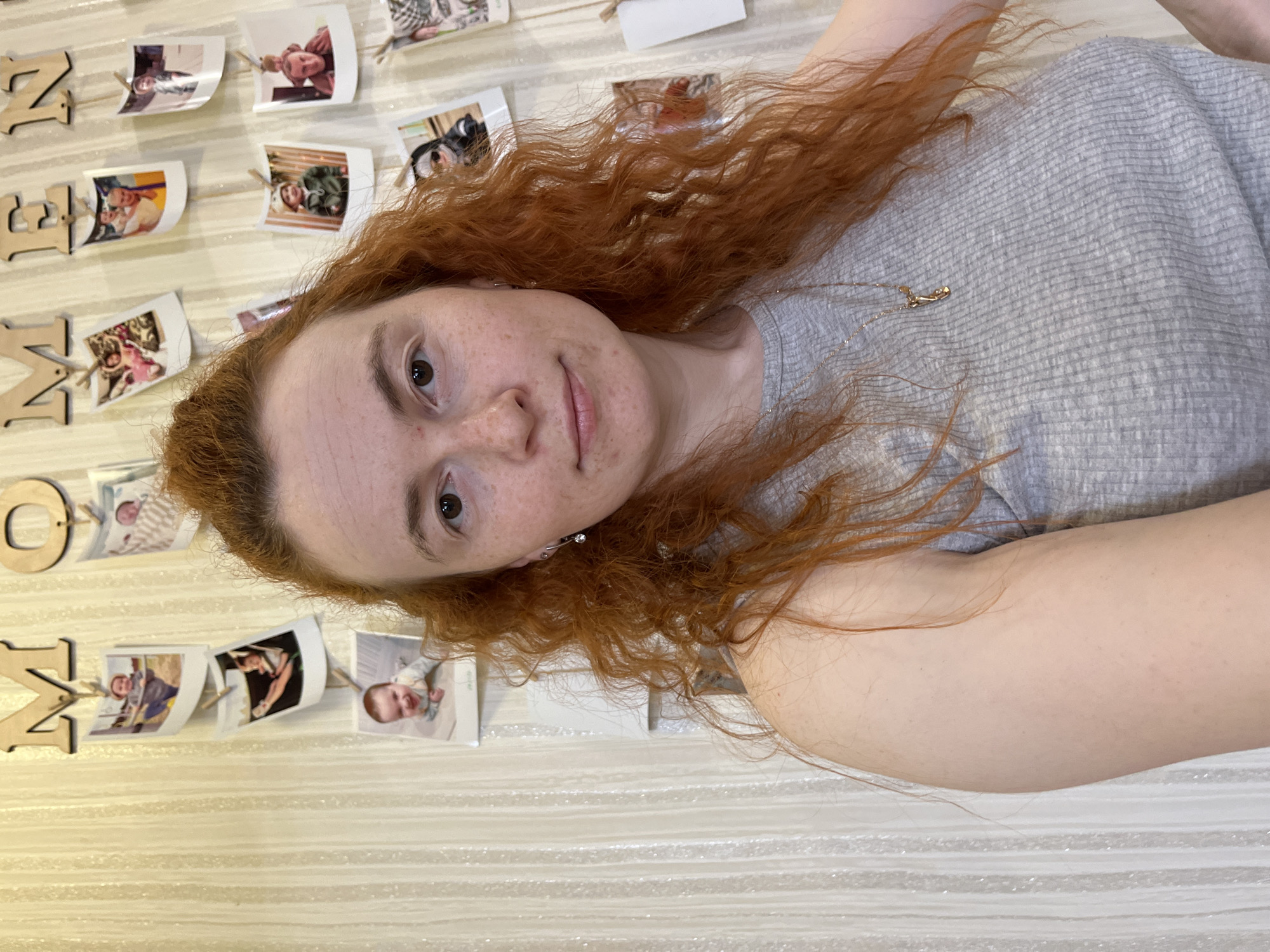 A selfie depicts a fair-skinned woman with red, curly hair and freckles. She is wearing a light grey, ribbed top and a delicate gold necklace. The background features a wall decorated with a "Z NAM RODZENSTWO" sign made of wooden letters and a string of photographs attached with clothespins. The setting appears to be inside a home. While the location may be within the city of Chaikovsky, Russia, this cannot be confirmed from the image itself.