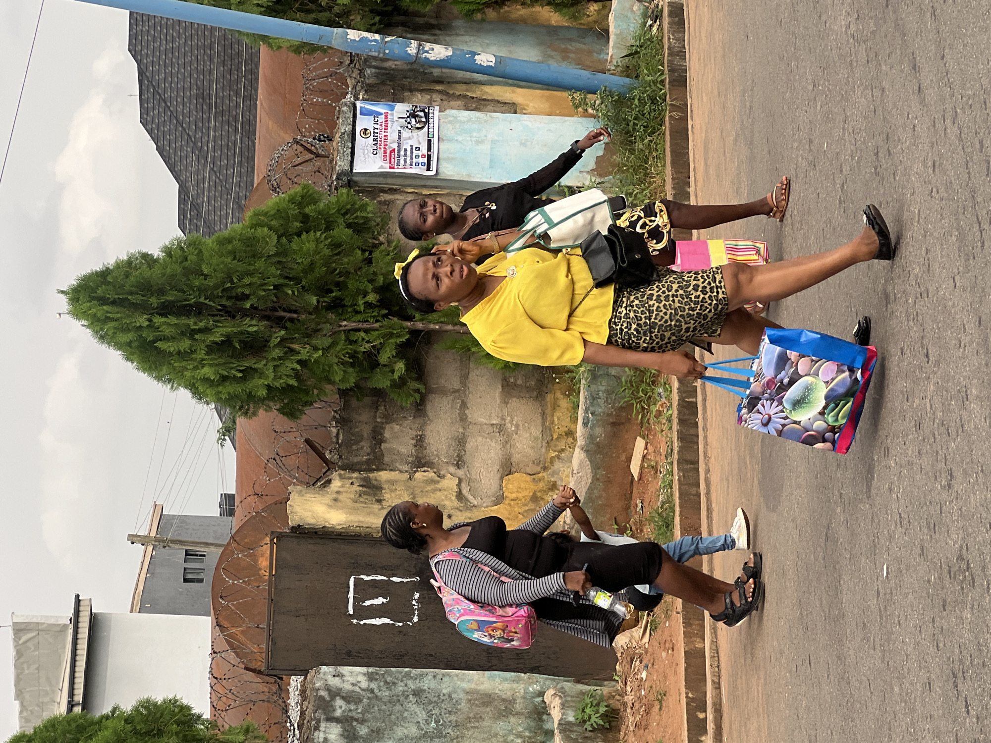 A medium shot captures three individuals walking along a paved road in Okpanam, Nigeria. In the foreground, a woman wearing a yellow blouse and a leopard-print skirt walks towards the right, carrying a shopping bag in her left hand and speaking on her cell phone. She has a black crossbody bag over her right shoulder. Behind her, a woman in a black top and patterned skirt walks, also towards the right, carrying a white and green tote bag. To the left of the foreground woman, another woman in a black dress and a striped cardigan walks away from the viewer, carrying a colorful backpack and a plastic bottle of water. A child, partially obscured, walks beside her, holding her hand and wearing blue jeans and white sneakers. The background features a low, weathered wall topped with barbed wire, behind which are various buildings and trees. A blue pole stands to the right of the frame. The sky is overcast.