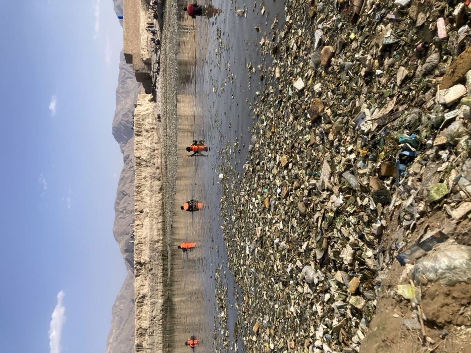 The image displays a daylight scene of a severely polluted waterway in Quetta City Tehsil, Pakistan. The foreground is dominated by a dense, heterogeneous accumulation of solid waste, including plastics, foam fragments, fabric, and construction debris, covering the shallow water or immediate bank. Within the murky, refuse-laden water, five individuals are visible. Four are wearing bright orange personal flotation devices and are partially submerged, appearing to be waist to chest deep. One individual on the far right is similarly submerged, wearing a dark red garment. Some individuals are holding long poles, and a rope extends horizontally across the water, connecting several figures, suggesting a coordinated effort, likely for waste removal or retrieval. In the mid-background, a prominent, light-brown earthen embankment stretches horizontally, with numerous small figures (people) observed standing atop it. To the far right, behind the embankment, a brown masonry structure with a blue container is visible. The distant background features a range of barren, light-brown mountains under a clear blue sky with scattered small white clouds.