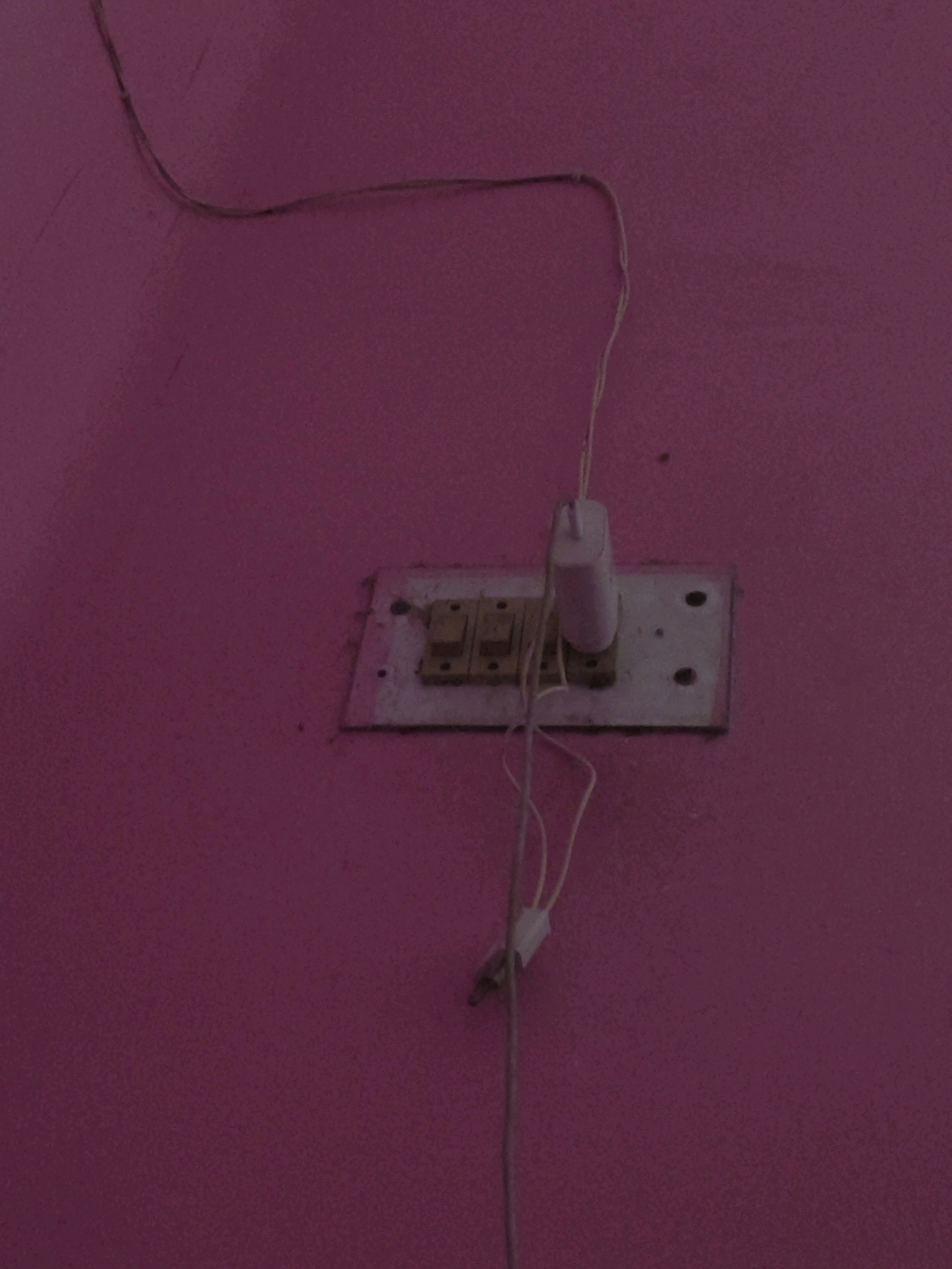 The image shows a close-up of a pink wall in Khillupur, India. A white electrical socket with two switches and a white mobile phone charger plugged into one of the sockets is mounted on the wall. Two wires are visible, one running from the top left of the frame and another connected to the charger. The overall scene is dimly lit, with the pink wall appearing somewhat desaturated. There are no people or other objects visible in the frame.