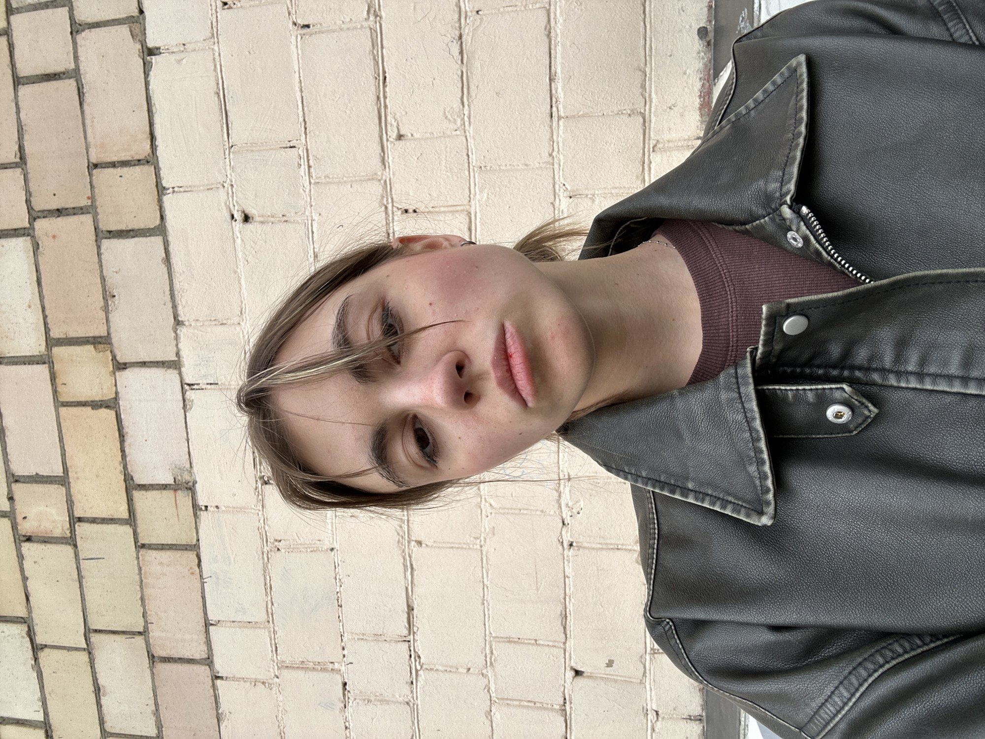 A young woman with light brown hair pulled back in a ponytail and bangs is facing the camera. She has a neutral expression and is wearing a dark jacket over a burgundy shirt. The background is a light-colored brick wall. There are no discernible Moscow landmarks or city-specific elements present in the image.