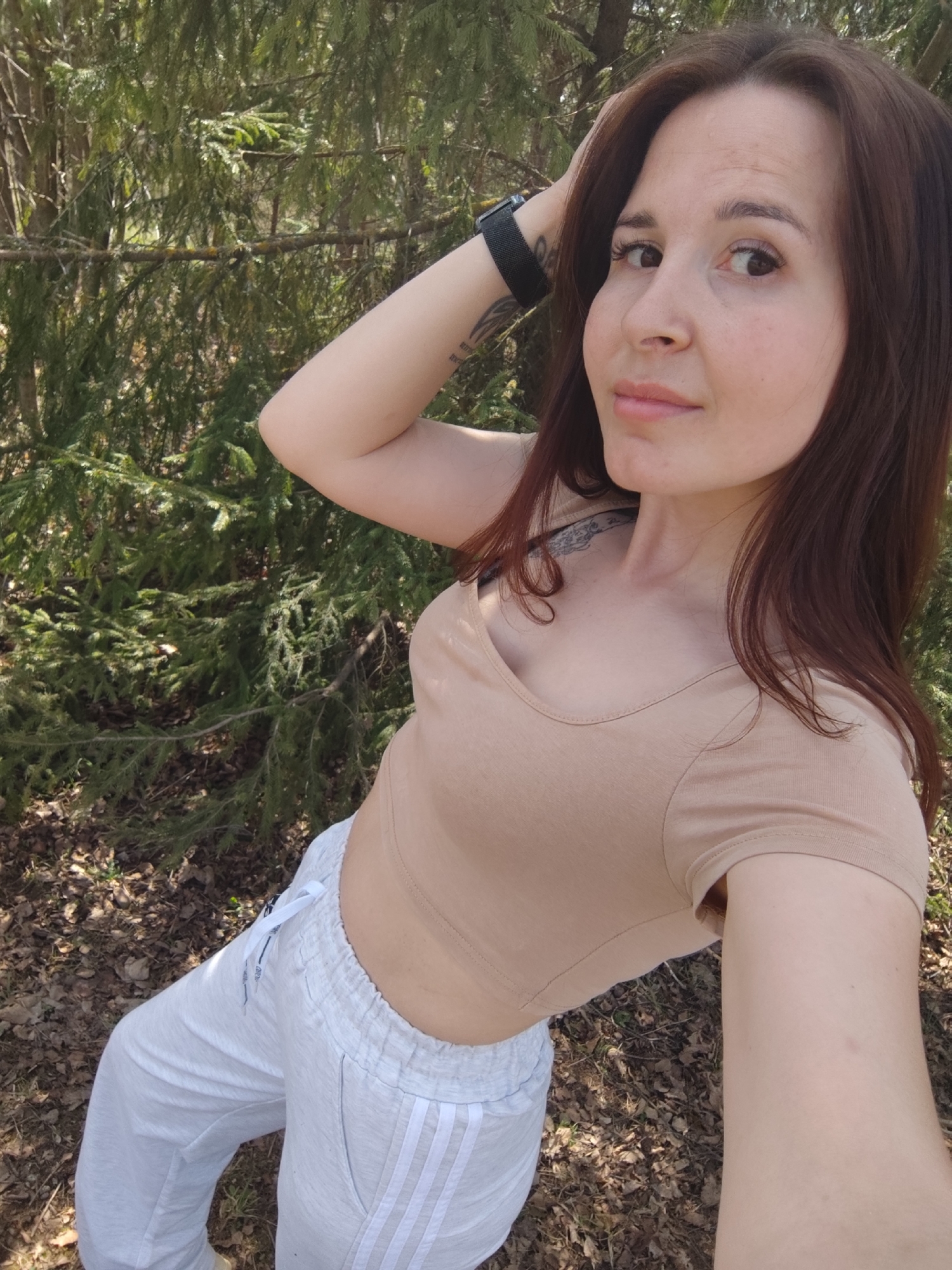 A woman is in a wooded area, seemingly taking a selfie. She is wearing a light-colored crop top and white athletic pants with three stripes down the side. She has brown hair, and a wristwatch is visible on her left wrist. The background shows green trees and leaf-covered ground, indicative of a natural or park setting. No evidence in the photograph indicates that the photograph was taken in the city of Фоки, Russia.