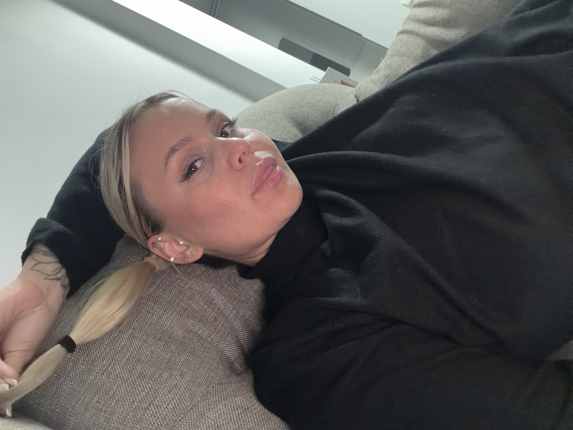 A woman with blonde hair in a ponytail is lying on a couch. She is wearing a black turtleneck sweater. Her left arm is visible, and she has tattoos on her forearm. She has multiple piercings in her left ear. The background includes a white wall and a light fixture. There is no information in the image to suggest that the location is in the city of Sarapul, Russia.