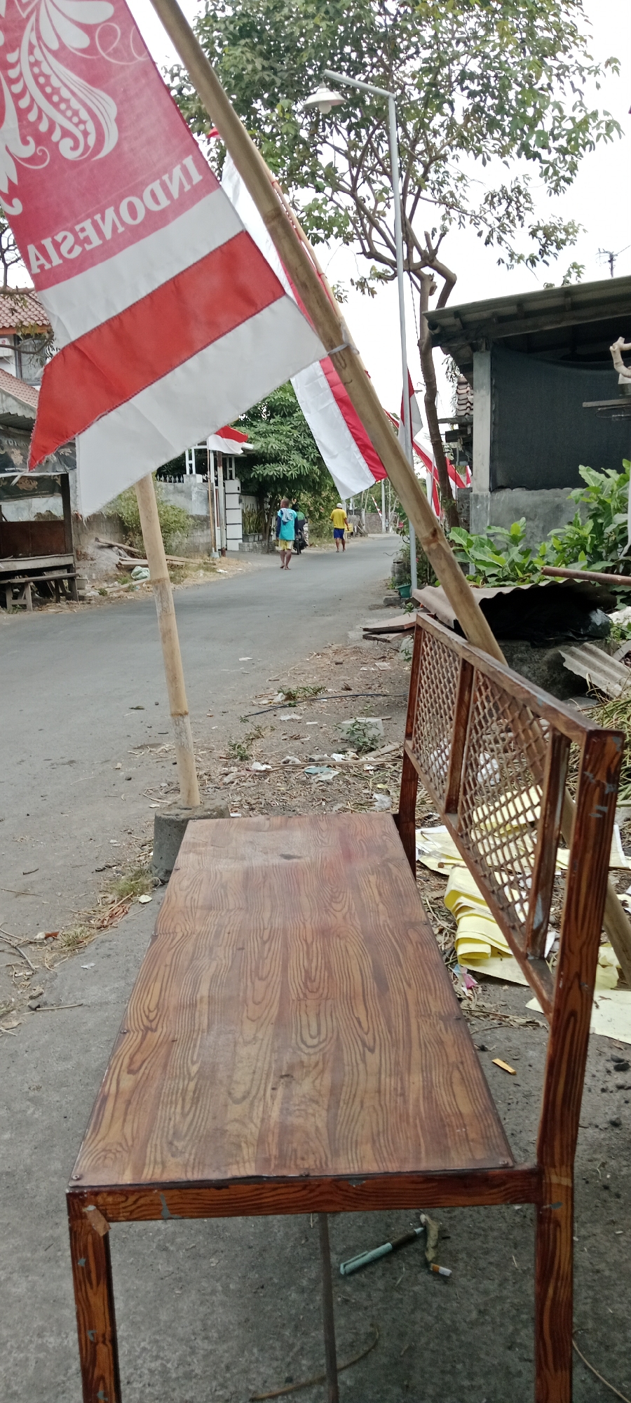 In Purwomartani, Indonesia, a wooden bench with a patterned seat and a woven backrest is positioned on a paved street. Two Indonesian flags, red and white, are prominently displayed on bamboo poles, one of which is leaning against the bench. In the background, two men are walking down the street, one wearing a blue t-shirt and yellow shorts, and the other in a yellow shirt and shorts. A street lamp with a white fixture stands near a leafy tree. The environment appears to be a residential area with houses and some scattered debris on the roadside.