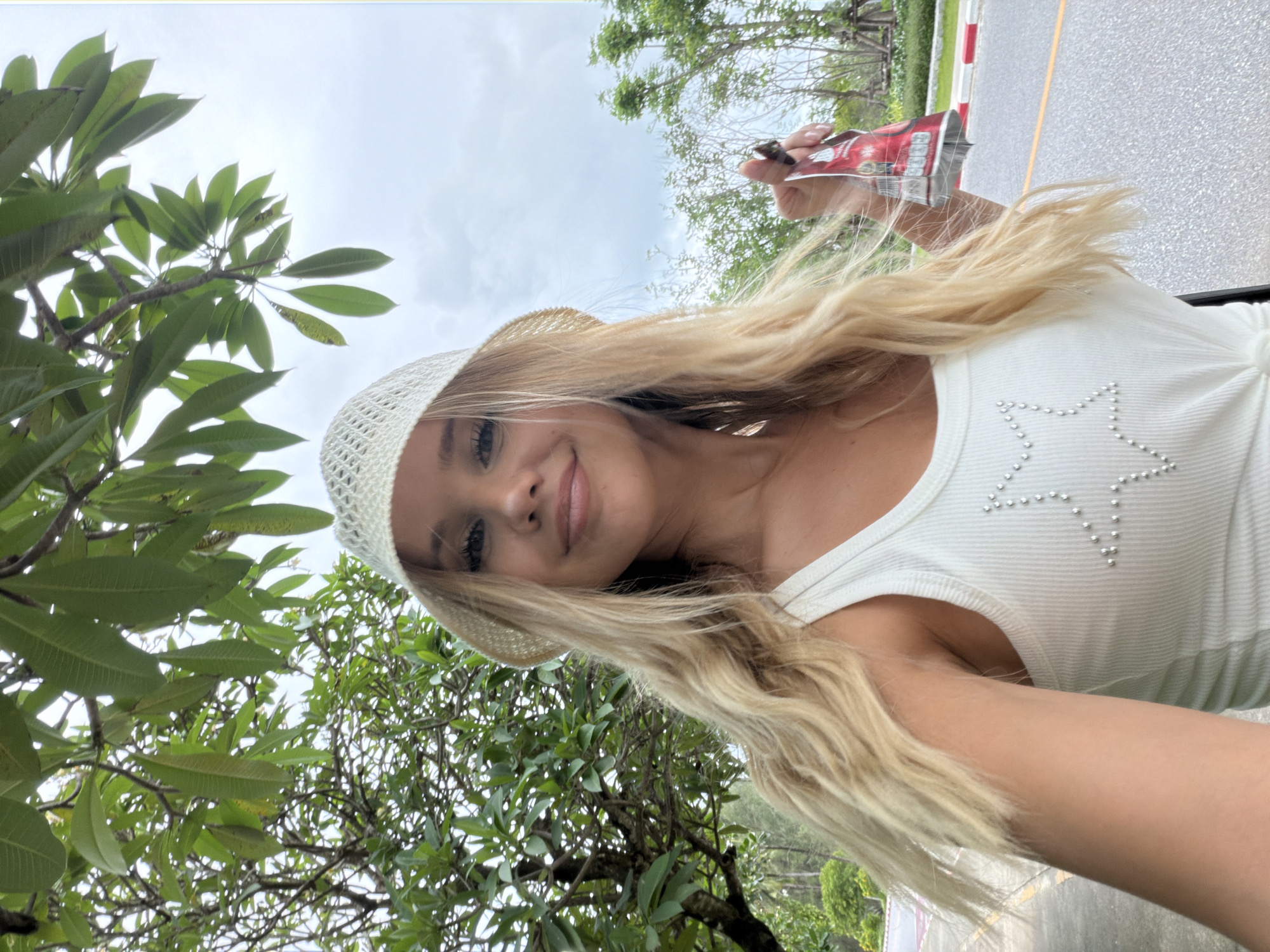 In Choeng Thale, Thailand, a young Caucasian woman with long, wavy blonde hair and a white knitted bucket hat is captured in a selfie. She is wearing a white ribbed tank top adorned with a silver star made of small studs. Her right hand, in the foreground, holds a partially opened red foil packet. The background features lush green foliage from a large tree overhead, a paved road with yellow and white markings, and a cloudy sky. The overall environment suggests a tropical, outdoor setting.