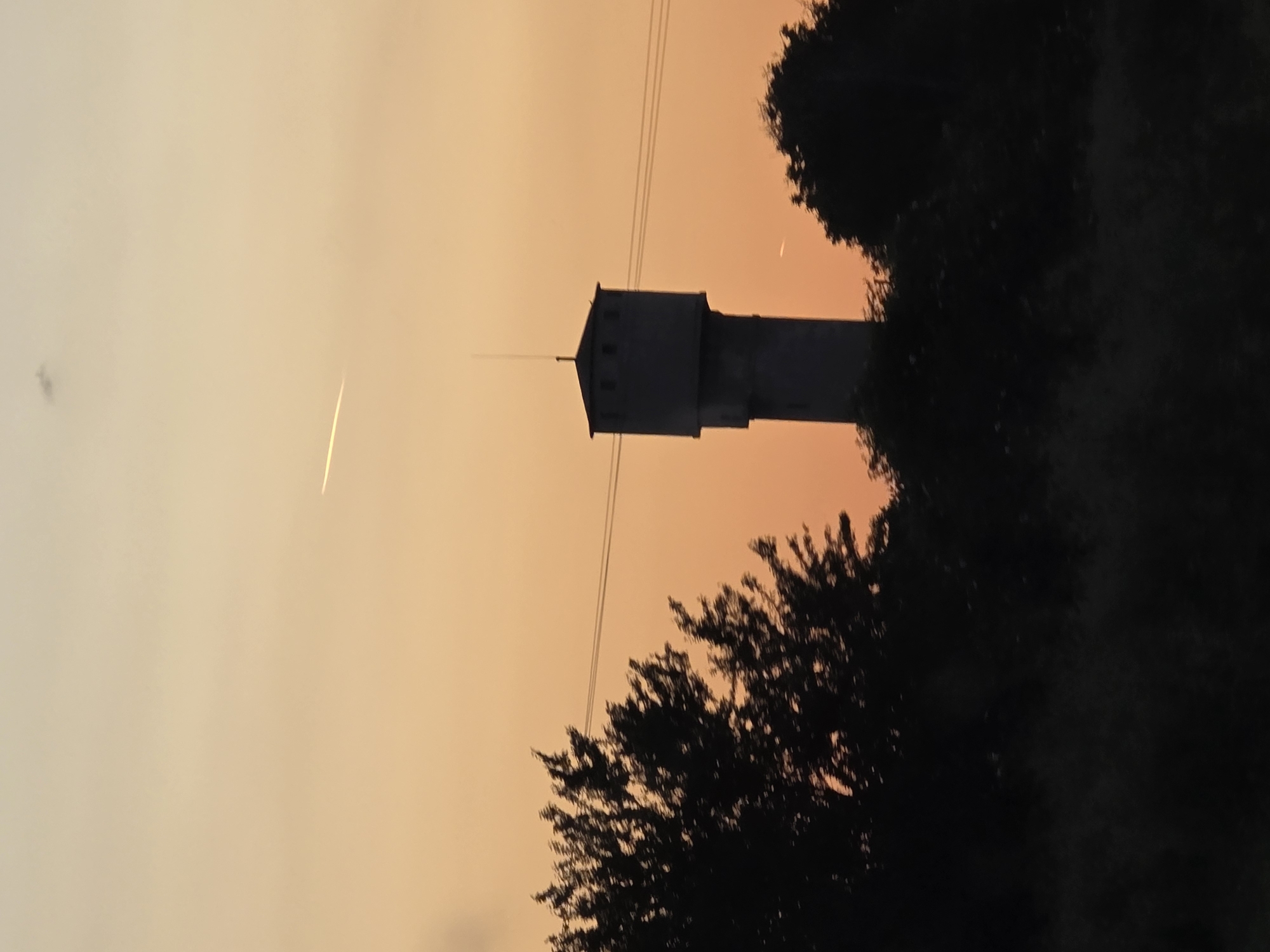 A landscape photograph in portrait orientation depicts a water tower silhouette in Karadjovo, Bulgaria, viewed against an orange-tinted sky at twilight. Darkened foliage frames the tower at the base and partially obscures its right side. Power lines run across the sky. An airplane contrail appears in the sky to the left of the tower.