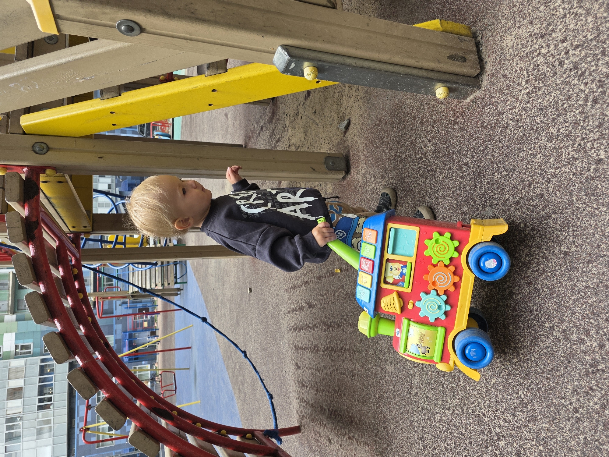 A young Caucasian child with blonde hair is on a playground, pulling a colorful toy train. The child is wearing a dark sweatshirt and patterned shorts. The toy train is red, yellow, and blue with various interactive features. The playground has a soft, dark brown, granular surface. In the background, there is a large modern residential building and other playground equipment, including a red archway and yellow and brown wooden structures. The scene appears to be outdoors in an urban environment.