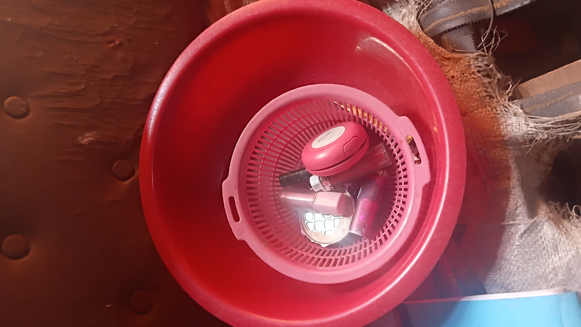 A red plastic basin contains a smaller pink plastic colander, which holds several cosmetic items. These include a compact mirror, a pink lipstick or gloss tube, and a pink compact makeup case. The basin and colander are placed on a textured surface that appears to be burlap or a similar woven material, partially obscuring a blue object to the left and a dark brown textured background behind. This scene is located in the city of Prabumulih, Indonesia.