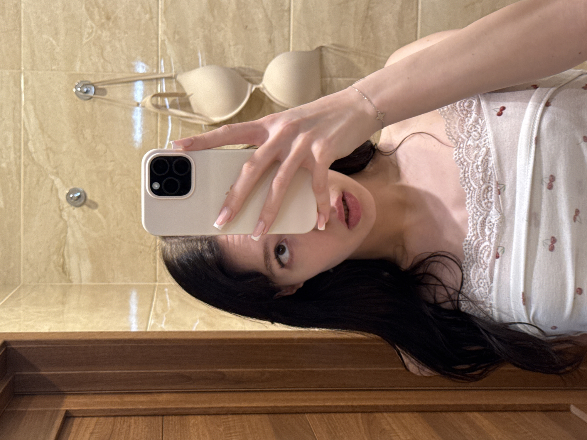 In a bathroom with beige marble tiles and a wooden door, a young woman with long dark hair takes a selfie in a mirror. She is wearing a white camisole with a cherry pattern and a lace trim. Her right hand holds a smartphone, partially obscuring her face. A beige bra is hanging on hooks on the tiled wall behind her.