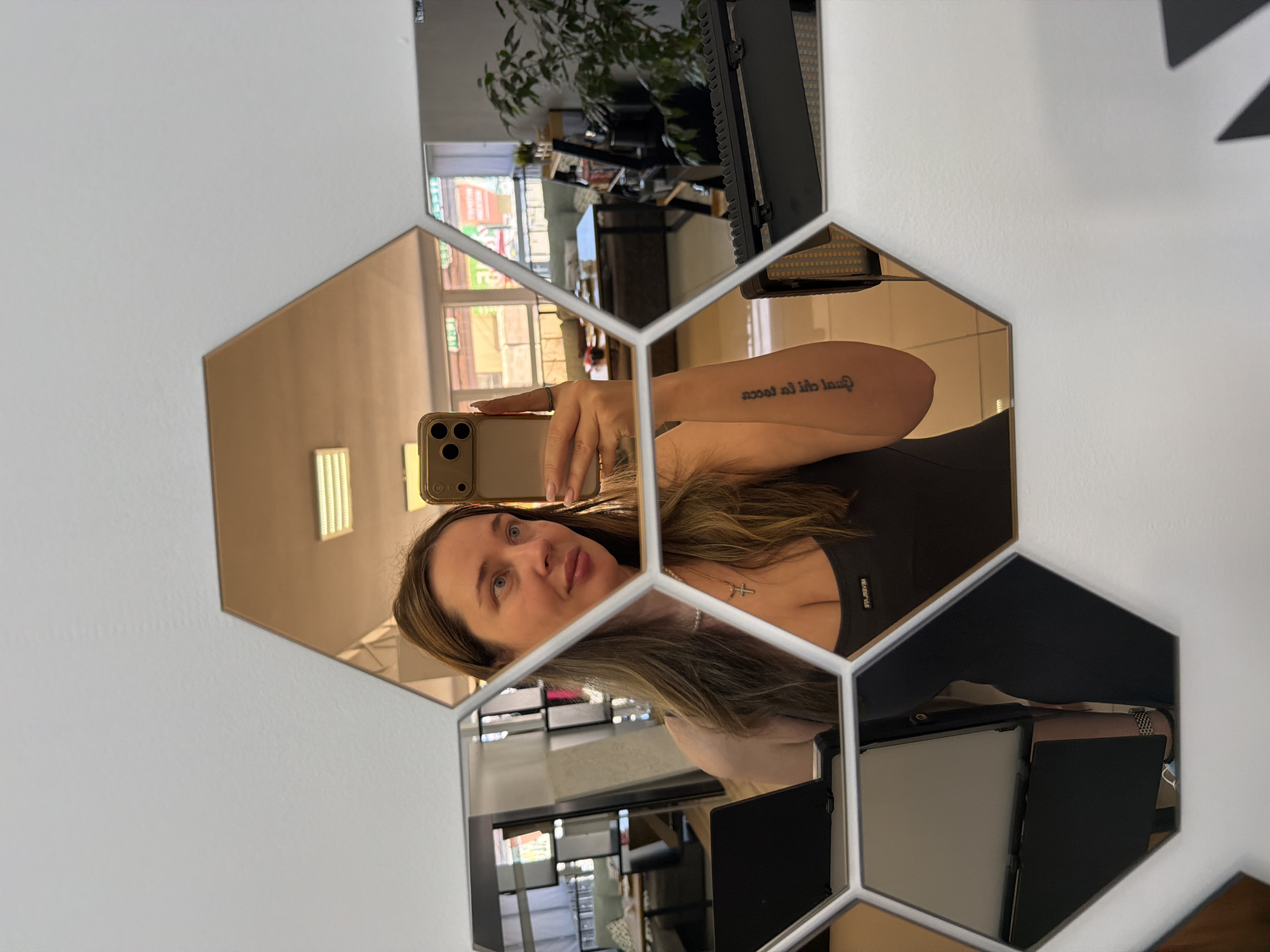 A female subject is depicted in a selfie taken in a multi-segment hexagonal mirror array mounted on a white wall. The subject, a Caucasian woman with long brown hair and blue eyes, looks slightly upward and to her left, holding a light-colored smartphone with a triple-lens camera system in her right hand. She wears a dark grey tank top with "NEVERLATE" printed on the upper chest, a pearl necklace with a silver cross pendant, and has a tattoo with text on her left forearm. Her fingernails are painted light pink.
The reflections in the mirror indicate an indoor commercial environment located in Chaikovsky, Russia. To the subject's left, dark furniture, including seating and tables, and a multi-tiered shelving unit are visible. To her right, a large window reflects an exterior street scene featuring building facades and multiple commercial signs with Cyrillic text, including "ОБОИ" (Wallpaper), "РЫНОК" (Market), and elements suggestive of "ТЕЛЕ2" (Tele2, a Russian mobile operator). Green foliage from outdoor trees or shrubs is also reflected. The interior reflections also show a large potted plant, additional tables and seating, and a rectangular ceiling light fixture. In the lower right foreground, a black electronic device with a panel of amber LEDs, consistent with a portable photographic light, is partially visible.
