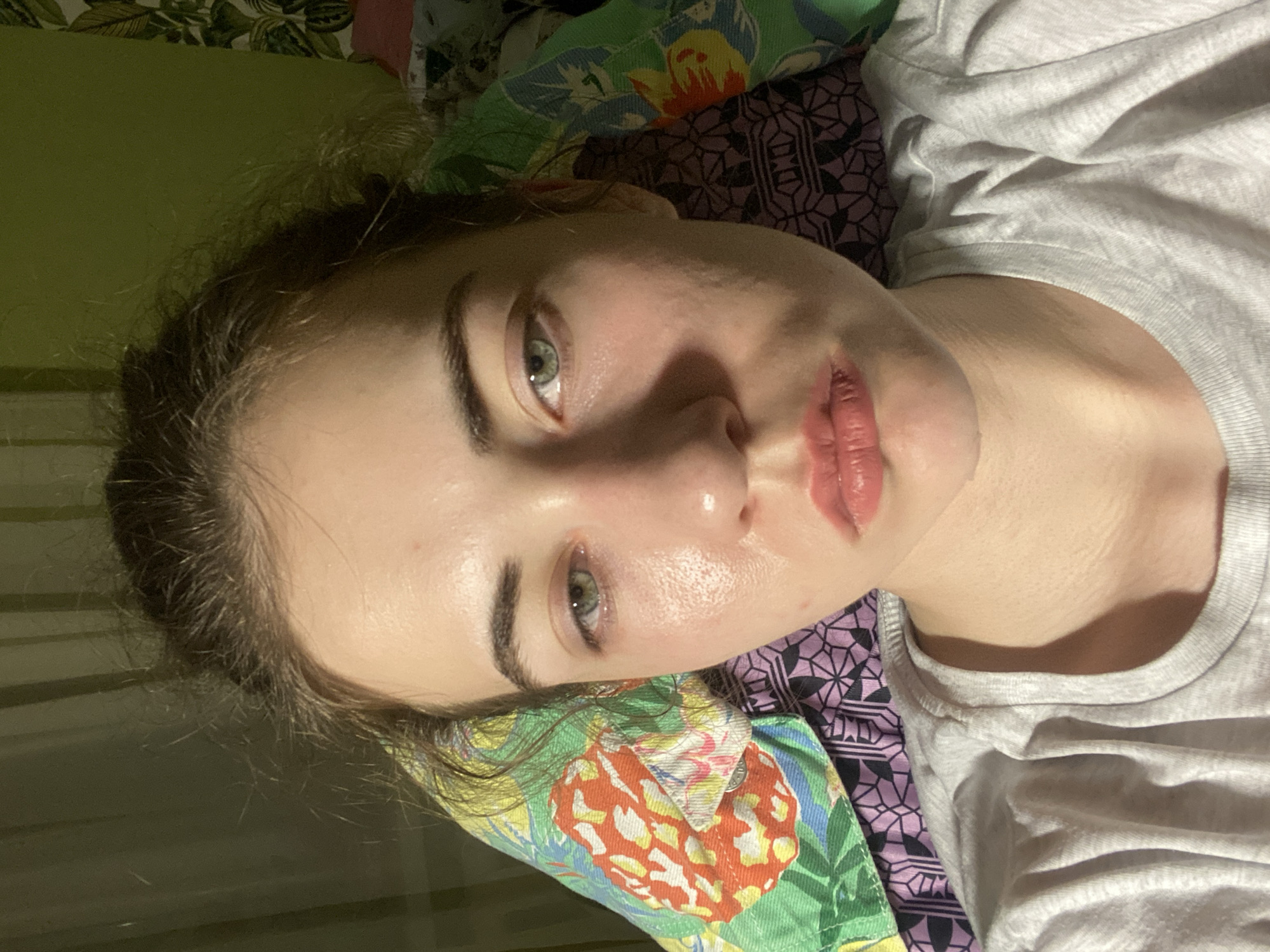 The media file shows a close-up, overhead shot of a young person lying down. They are wearing a light gray t-shirt and resting their head on a pillow with a vibrant, tropical pattern featuring large red and yellow floral designs and green leaves. The background consists of a plain green surface and patterned bedding.
The person has fair skin with a dewy sheen, suggesting it might be morning or that they have just applied skincare products. Their eyes are open and gazing upwards, with one eye more visible than the other. The visible eye is a striking green with dark eyebrows. Their lips are slightly parted and appear to have a glossy finish, possibly from lip balm or gloss. Their brown hair is loose and frames their face.
The lighting in the image appears to be artificial and somewhat dim, casting shadows and highlighting the texture of the skin. This, combined with the person lying down, suggests a relaxed or possibly even sleepy state. The setting seems to be indoors, likely a bedroom, given the presence of bedding and a pillow. There is no visible text in the image. The overall mood is intimate and candid.