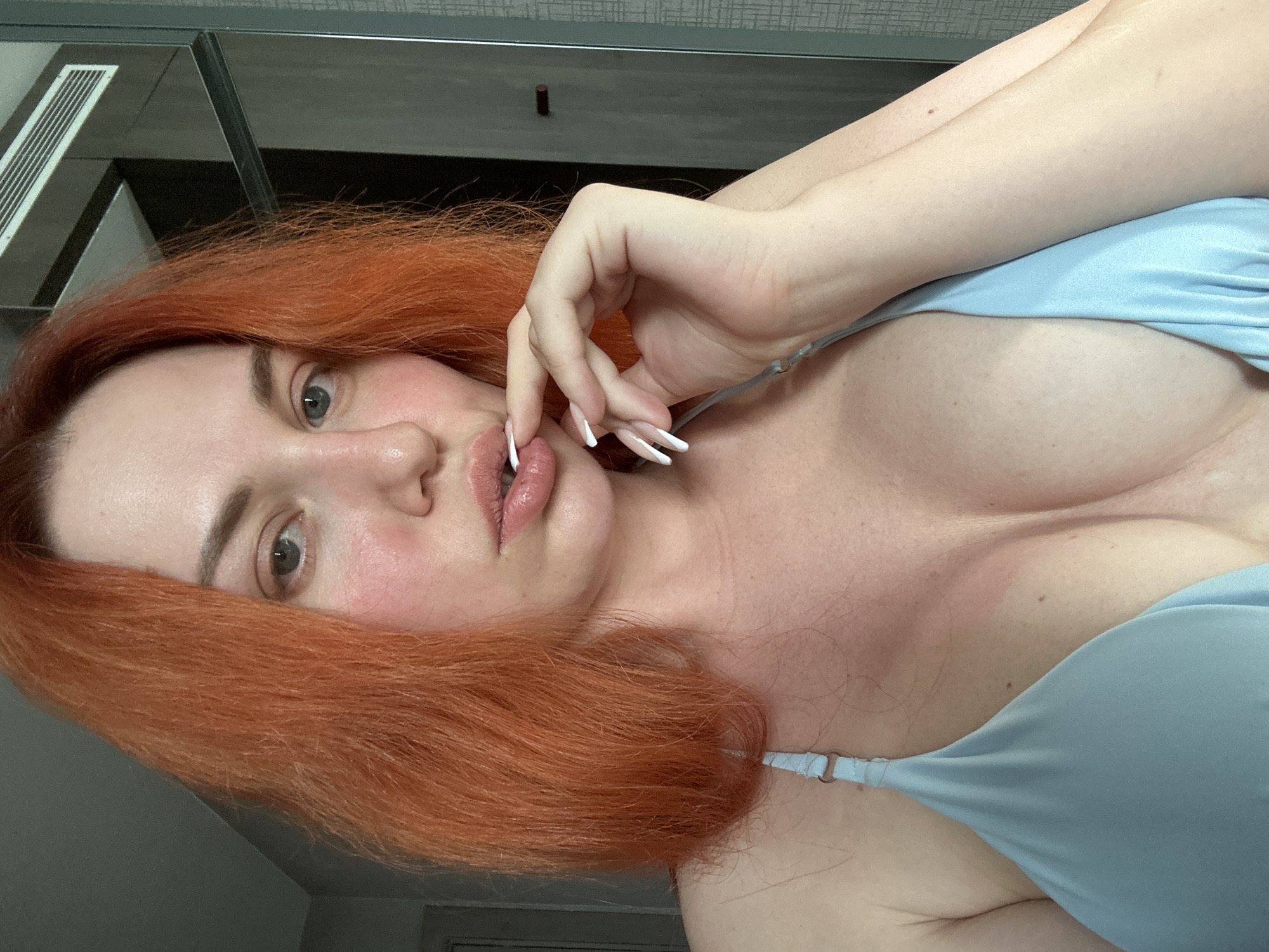 The image shows a close-up of a woman with bright orange hair. She is wearing a light blue halter top and has her hand near her mouth. Her fingernails are long and white. Her skin appears flushed, and there is a visible tan line on her chest and shoulder. The background is blurred but appears to be an indoor setting with a mirrored surface and a vent visible. The context of Avcılar, Turkey is not present in the image.