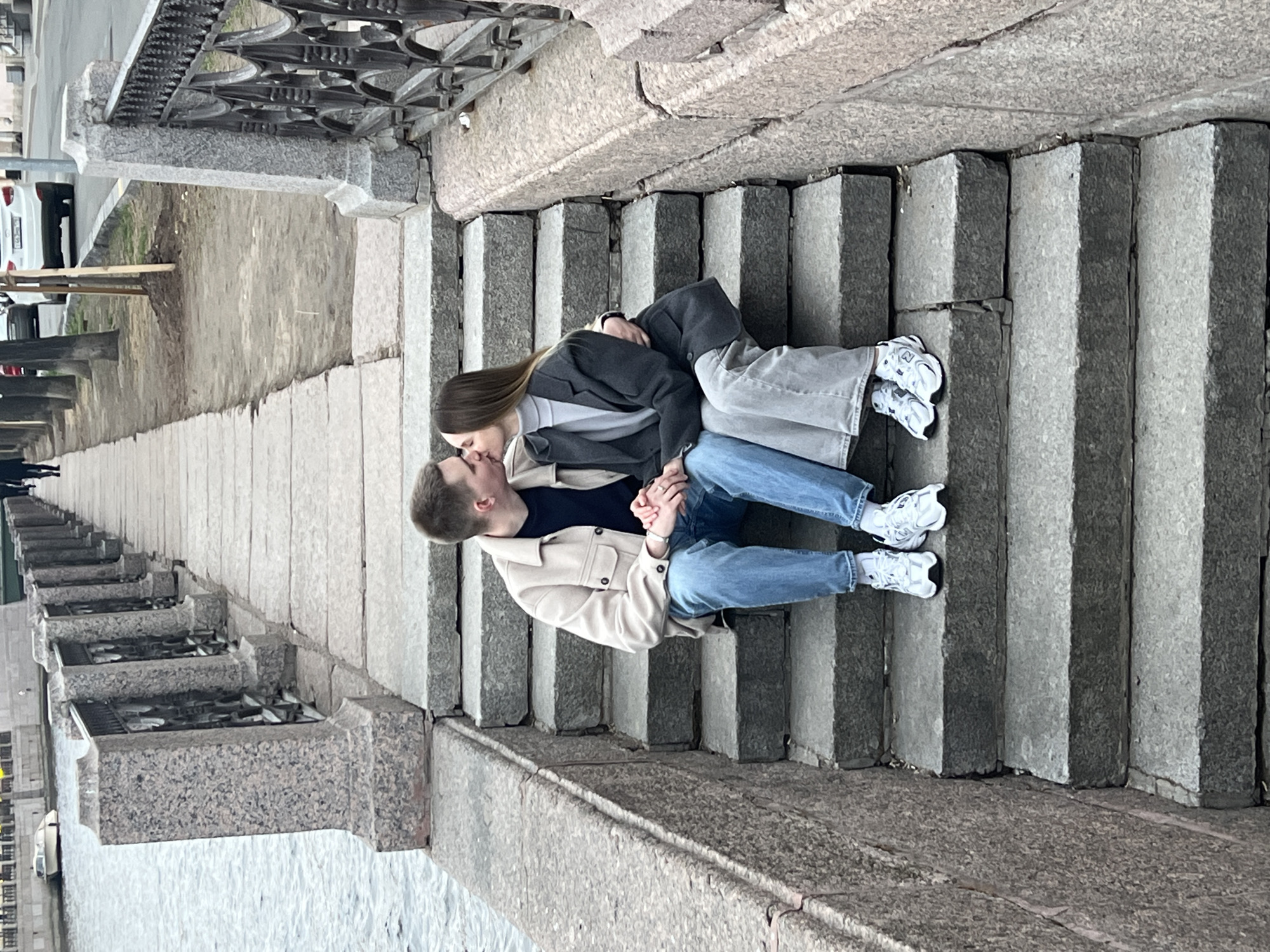 A man and a woman are kissing on a stone staircase in Saint Petersburg, Russia. The couple is sitting on the stairs, with the woman on top of the man. The woman has long brown hair and is wearing a gray blazer and gray jeans. The man is wearing a beige jacket, a dark blue shirt and blue jeans. They are holding hands. The background shows the canal and a stone bridge. The photo seems to be taken from a high angle.