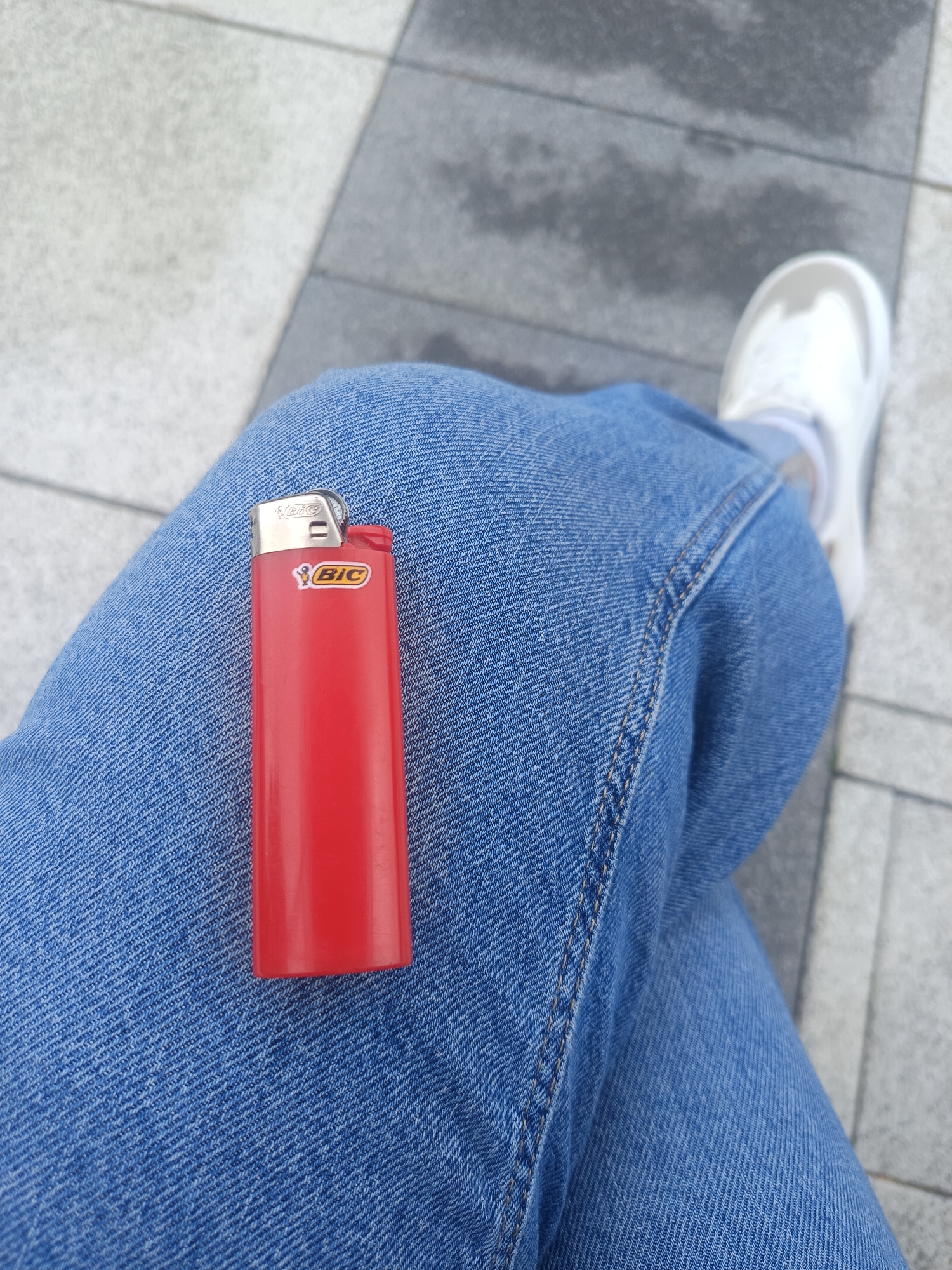 A close-up, eye-level shot shows a person's legs dressed in blue denim jeans and a white sneaker. A red BIC lighter is placed on the jeans, near the person's thigh. The background consists of a sidewalk made of gray and light-colored rectangular paving stones, suggesting an outdoor urban setting. The lighting indicates it is daytime. There is no specific information in the image to determine the location within Świnoujście, Poland.