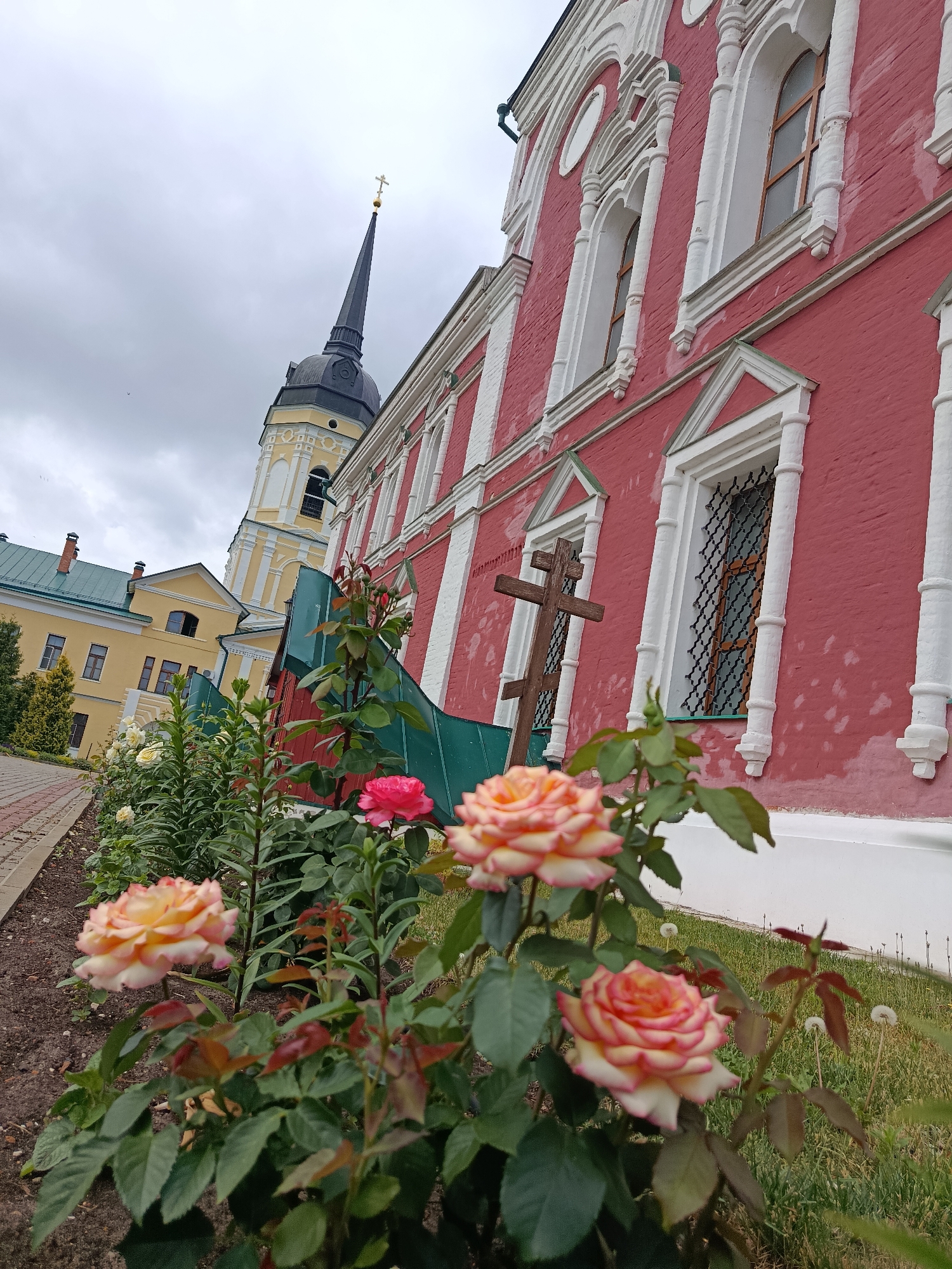 The image shows a view of the Holy Trinity Church in Radovitsy, Russia, from a garden in front of it. In the foreground, several roses are in bloom, with a few buds and leaves visible. Behind the roses, a wooden cross stands planted in the ground. The church itself is a large, pink building with white trim around its windows and cornices. A tall, slender bell tower with a black spire topped by a golden cross rises behind the church. To the left of the church, another building with a green roof is visible, also appearing to be part of the religious complex. The sky is overcast and gray, suggesting a cloudy day. The ground in the foreground is covered with soil and grass. A paved walkway runs along the left side of the image.
