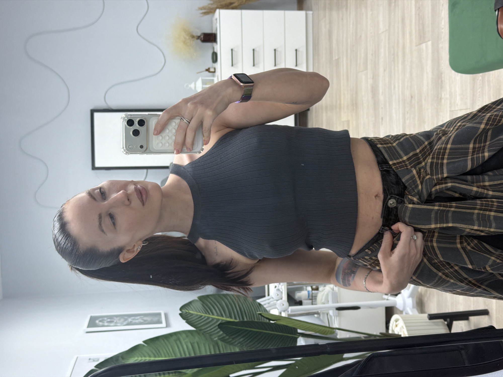 A woman with dark hair pulled back into a ponytail, wearing a grey ribbed crop top and a plaid flannel tied around her waist, is taking a selfie indoors. She is holding a cell phone in her right hand, which has a clear case and a white grip. Her left hand is near the waistband of her pants. She is wearing a watch on her right wrist and rings on both hands.
The background includes a white dresser with six drawers, a framed black and white picture on the wall, and a green chair. There is also a potted plant with large green leaves in the foreground. The scene appears to be in a room with light-colored walls and flooring. There is no evidence in the photo to suggest that this image was taken in Perm, Russia.
