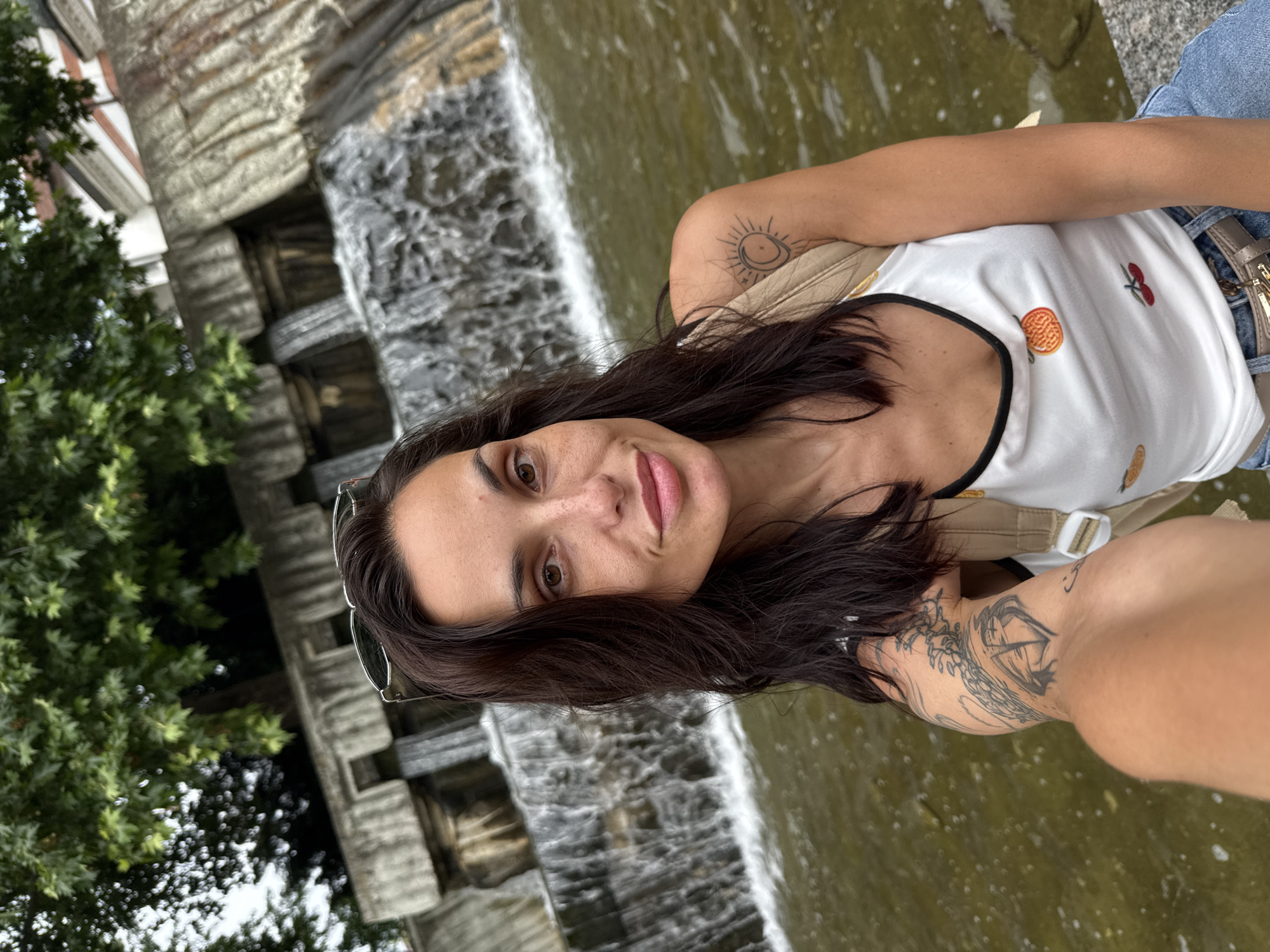 The image displays a selfie featuring a woman in front of the artificial Cascades waterfall in Kaylaka Park, Pleven, Bulgaria.
The subject is a woman with dark brown, shoulder-length hair, parted to her right. She has brown eyes and visible freckles on her nose and upper cheeks, exhibiting a slight smile with closed lips. She wears a white tank top with black trim, embroidered with fruit designs, specifically an orange and cherries. A beige backpack strap is visible over her right shoulder, and she wears light blue denim jeans with a light beige belt featuring a gold-toned metallic buckle. Sunglasses are positioned atop her head, partially obscured by her hair. She has two visible tattoos: a stylized sun and crescent moon within a circle on her right upper arm/shoulder, and an intricate, multi-element tattoo in black ink covering a portion of her left forearm and bicep.
The background is dominated by the multi-tiered Cascades waterfall, constructed from light-colored concrete or stone, with water actively flowing down its levels. This waterfall empties into a body of still, greenish-brown water. Above and around the waterfall structure, dense green deciduous trees with prominent leaves are visible, indicative of an outdoor park environment. The sky, observed through the tree canopy, appears overcast. In the upper right background, above the waterfall, architectural elements of a building, including a section of a reddish roof and a white facade with dark trim, are faintly discernible.