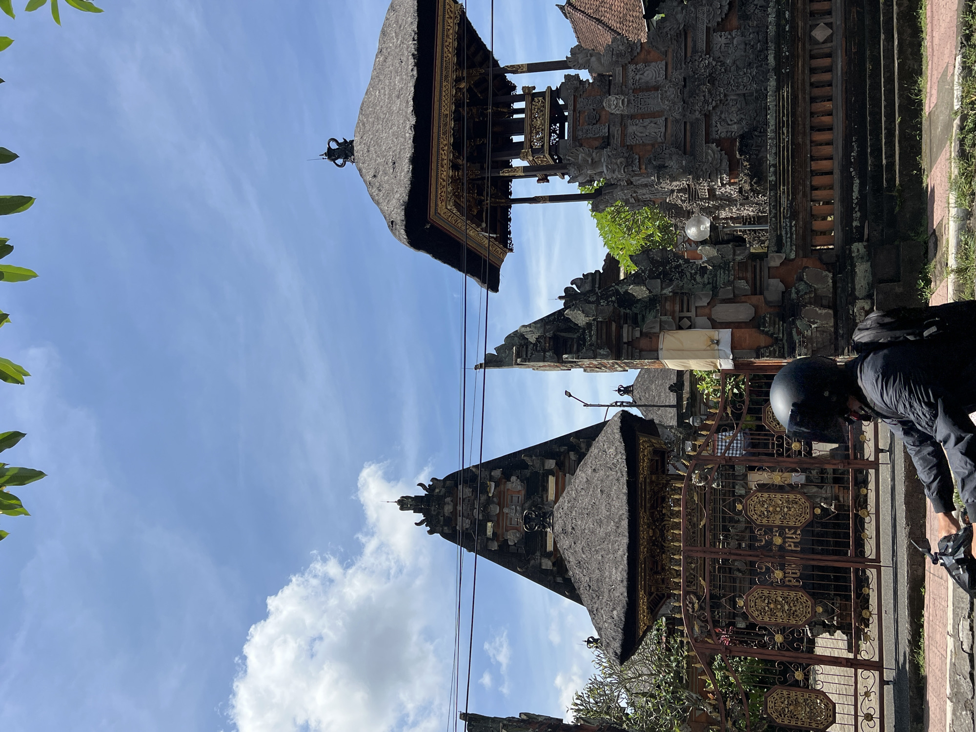 The image shows a street scene in Peliatan, Indonesia, with a traditional Balinese architectural structure prominently featured. The structure, likely a temple or shrine, has a tiered, thatched roof and ornate carvings. The sky is blue with scattered clouds. Electrical wires run across the view. A person wearing a black helmet and riding a scooter is on the right edge. A decorative metal gate is partially visible, with intricate gold-colored details.