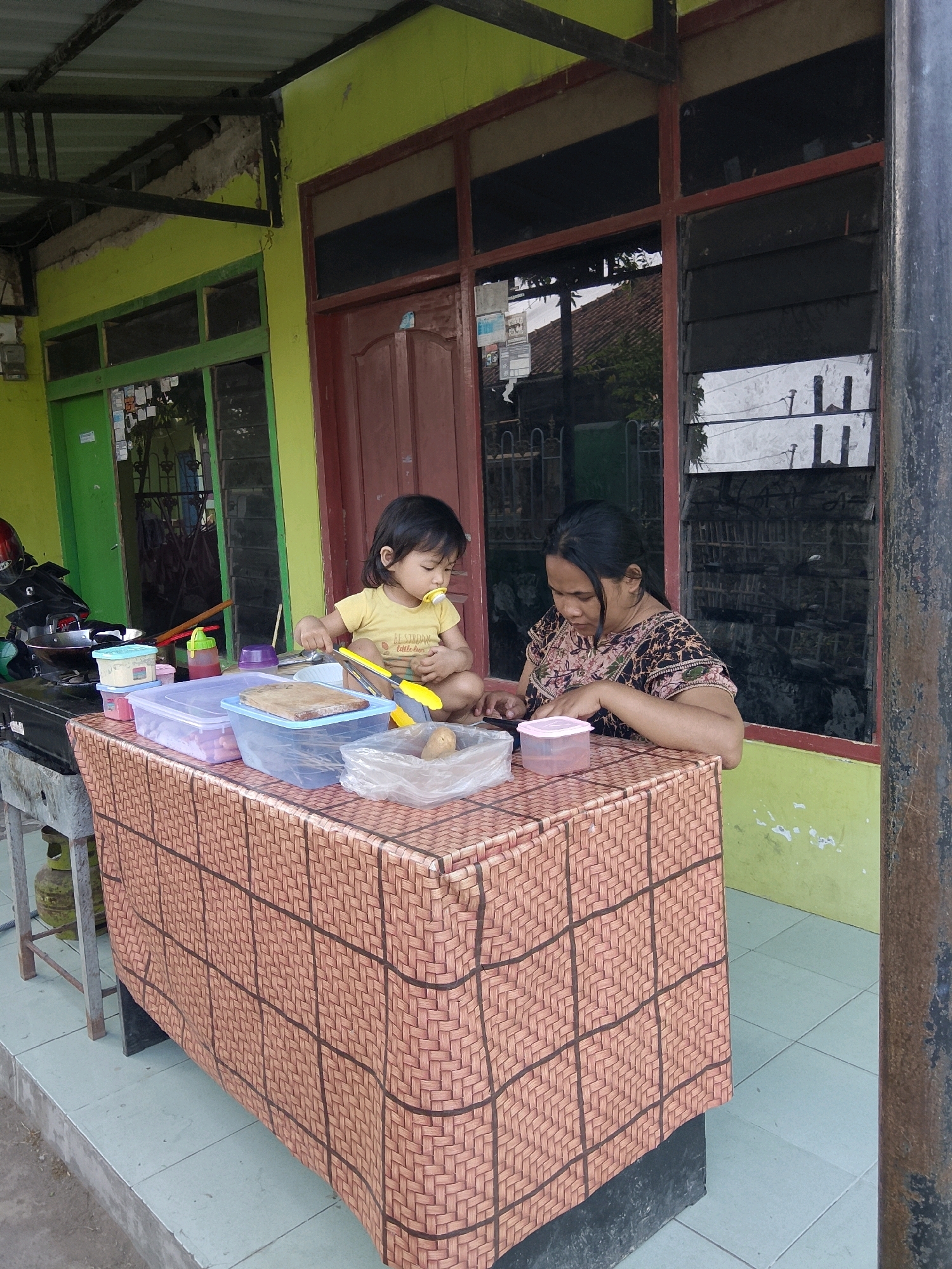 In Cirebon, Indonesia, a woman and a young child are positioned at a table covered with a brown woven-patterned tablecloth. The woman, seated on the right, wears a dark, patterned shirt and appears to be looking at her phone. The child, on the left, is wearing a yellow t-shirt and has a pacifier in their mouth. They are both engaged with items on the table. The table is laden with various containers, including clear plastic tubs filled with food items, a wooden cutting board, and a plastic bag containing a potato. To the left of the table, a black stove with a frying pan on it is visible. Behind the table, a building with lime-green walls and windows with red frames is present. A motorcycle is parked to the far left, partially obscured by the stove. The scene suggests an outdoor food preparation or selling area.