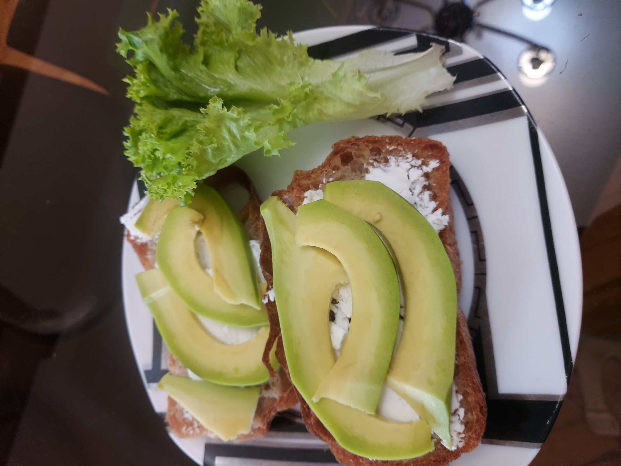 Two slices of toast topped with a white spread and sliced avocado are served on a plate with a lettuce leaf to the side. The plate has a black and white striped pattern on the rim. The food is arranged in a close-up shot, with the focus on the sandwiches and lettuce. The background is dark and out of focus. The scene appears to be a table setting, possibly for breakfast or a light meal. There is no indication of the location being in Yerevan, Armenia.