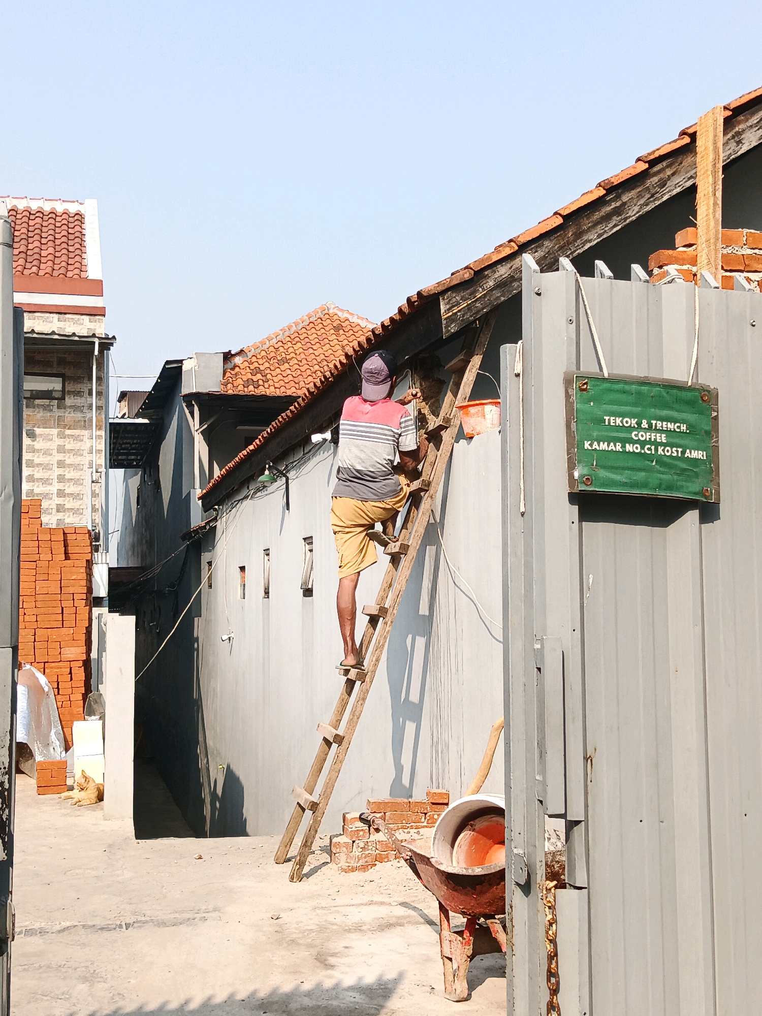 In a sunlit alleyway in Bandung City, Indonesia, a construction worker ascends a weathered wooden ladder against the side of a building. The worker, wearing a striped t-shirt and yellow shorts, is positioned on the ladder, facing away from the camera and towards the roofline. An orange bucket hangs from a nail near the top of the ladder, and an orange-handled tool rests on the roof. To the right of the worker, a grey corrugated metal fence displays a green sign with white lettering that reads "TEKOK & TRENCH. COFFEE KAMAR NO. C1 KOST AMRI". In the foreground, a red wheelbarrow with an orange basin filled with a reddish substance is visible. Stacked bricks are piled to the left of the scene, and a cat rests on the ground near them. The environment suggests an ongoing construction or renovation project, with exposed wooden beams and brickwork visible on the building's upper section. The sky is clear and blue.