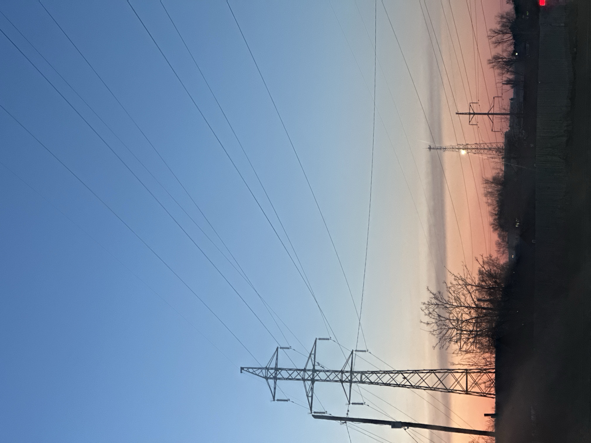 The image depicts a landscape under a sky transitioning from blue to pink, likely at dawn or dusk. Power lines stretch across the sky, connecting to transmission towers. The foreground is obscured by dark silhouettes of trees and possibly a road or embankment. In the distance, another transmission tower is faintly visible against the sky. Given the specified location of Chaikovsky, Russia, this is likely a rural or suburban area on the outskirts of the city with industrial infrastructure. No people or actions are visible in the image.