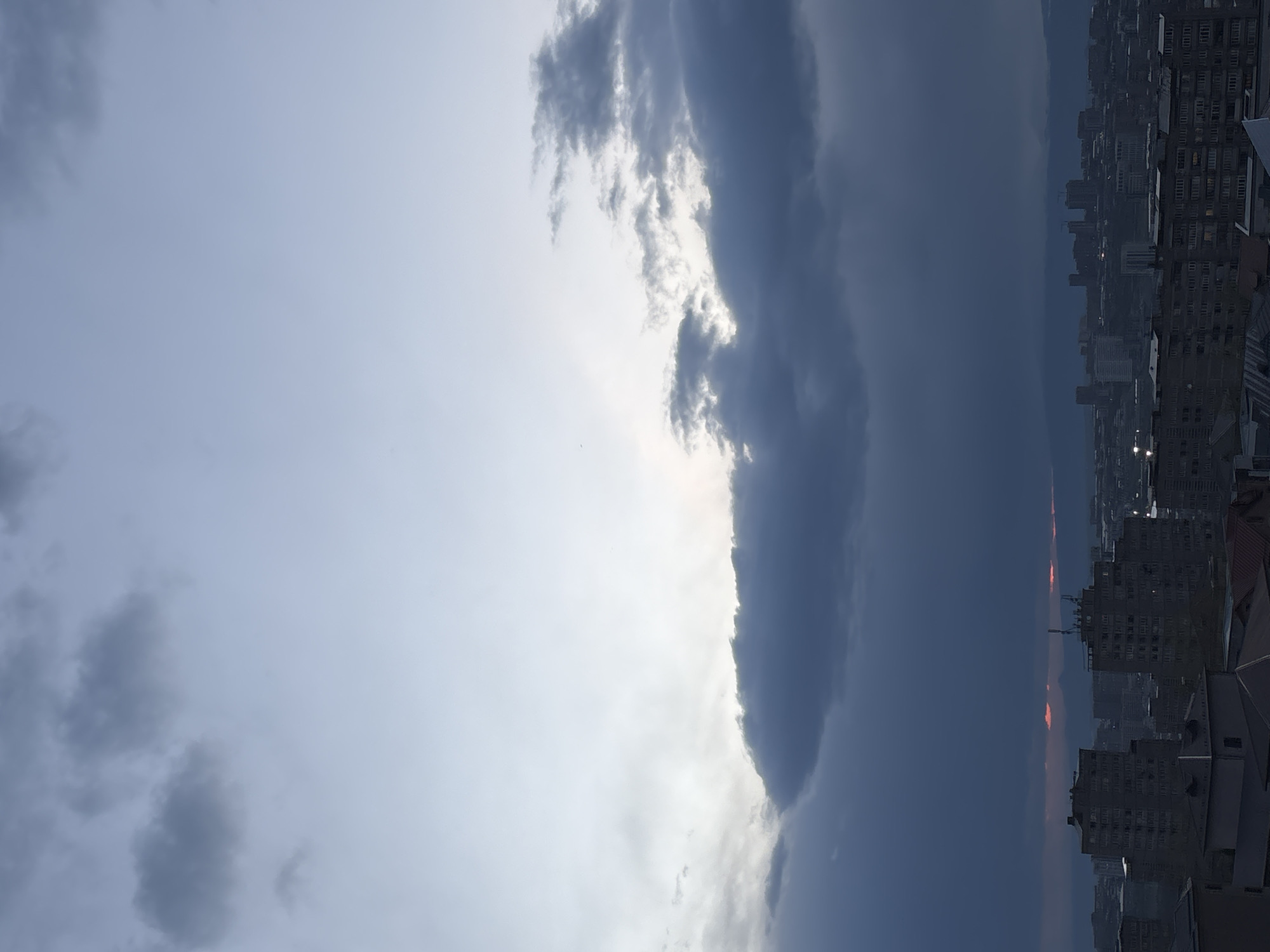 The image captures a dramatic urban landscape under a dynamic sky, viewed in a horizontal orientation. The scene is broadly divided by a striking weather pattern. The left half of the sky is predominantly bright and light blue-gray, with scattered, wispy clouds.
A prominent feature is a large, dense band of dark gray clouds stretching from the middle to the right side of the frame. The left edge of this cloud mass is intensely illuminated, possibly by direct sunlight, creating a bright "silver lining" effect against the darker cloud body. To the right of this bright edge, the sky transitions to a uniform, somber dark gray, suggesting heavy cloud cover or an approaching storm.
Below this turbulent sky, particularly on the right side of the image, lies a dense cityscape. Numerous multi-story buildings, appearing as residential or commercial structures, extend across the lower half of the frame towards the horizon. These buildings are largely in shadow, rendered in muted grays and browns, and appear somewhat silhouetted against the dark sky.
A narrow, faint band of reddish-orange light is visible along the distant horizon, positioned between the dark clouds and the buildings. This suggests the time of day is either sunrise or, more likely given the heavy clouds, late afternoon or early evening, marking a dramatic sunset. The overall lighting is characterized by strong contrasts between the bright, open sky and the dark, heavy cloud mass, casting the city below into shadow. The distant skyline hints at mountains or hills blending with the hazy horizon. The location context is Arinj, Armenia, adding a geographical reference to this atmospheric scene. There is no visible text in the image.