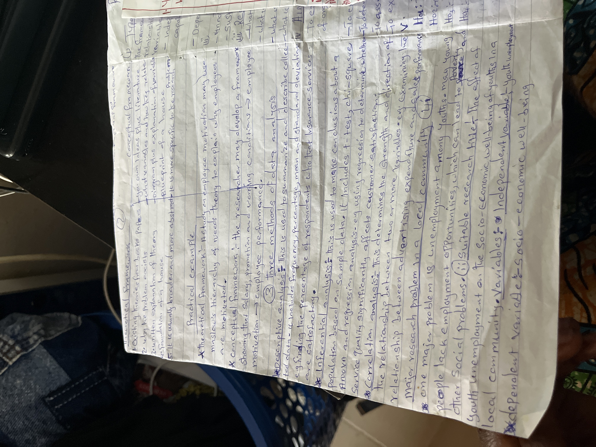 The image is a close-up, slightly angled shot of a notebook with handwritten notes in blue ink on lined paper. The notes appear to be academic, covering topics such as "Theoretical Framework," "Practical example," "Methods of data analysis," and "Correlation analysis." There are also sections on research problems and variables. The handwriting is in cursive and block letters. The notebook is placed on a dark, textured surface, possibly a table or desk. Part of a blue fabric, possibly clothing or a bag, is visible in the upper left corner of the image, along with some dark, shiny object that could be a phone or laptop. The overall lighting suggests an indoor environment, and the image is somewhat blurry, indicating movement or a lack of focus. There are no people or direct human interactions depicted in the frame. The context of Jos, Nigeria, is not visually evident in the image itself; this information appears to be provided externally.