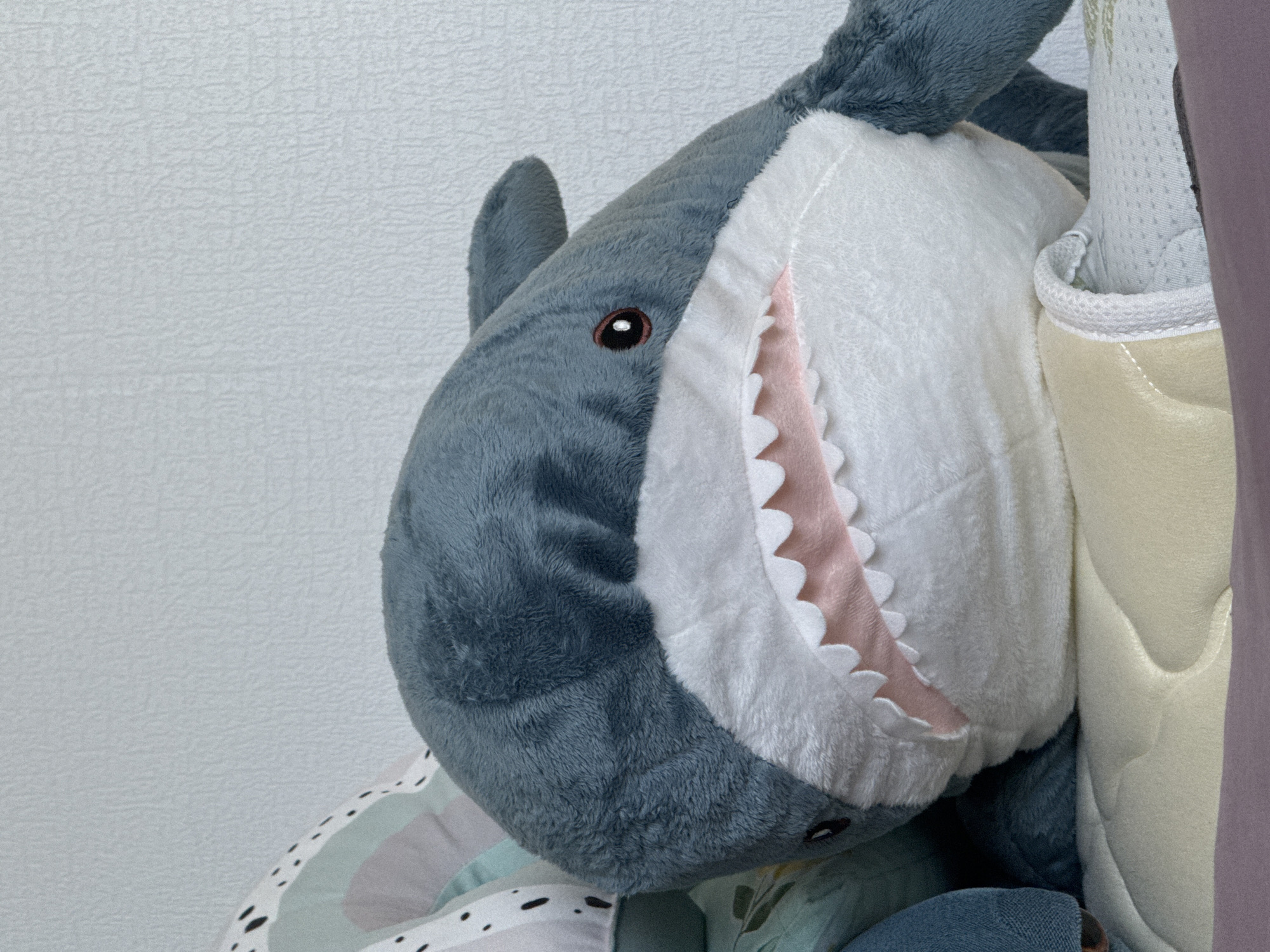 The image is a close-up, vertical, medium shot of a plush toy shark. The shark is gray with a white belly and pink interior of its mouth, which is agape, revealing rows of white teeth. It has a prominent dorsal fin and a visible eye with a dark pupil and an orange iris. The shark is positioned diagonally, with its head facing the right side of the frame, and it appears to be resting on a light yellow mattress or cushion. Behind the shark, to the left, a portion of another pillow with a rainbow and dotted pattern is visible. The background consists of a textured, pale blue or gray wall. The lighting is soft and diffused. There are no people or animals present in the image. The scene appears to be indoors, likely in a domestic setting such as a bedroom or playroom. The image does not contain any information or visual cues that suggest a location in Ankudinovka, Russia, or any other specific city.