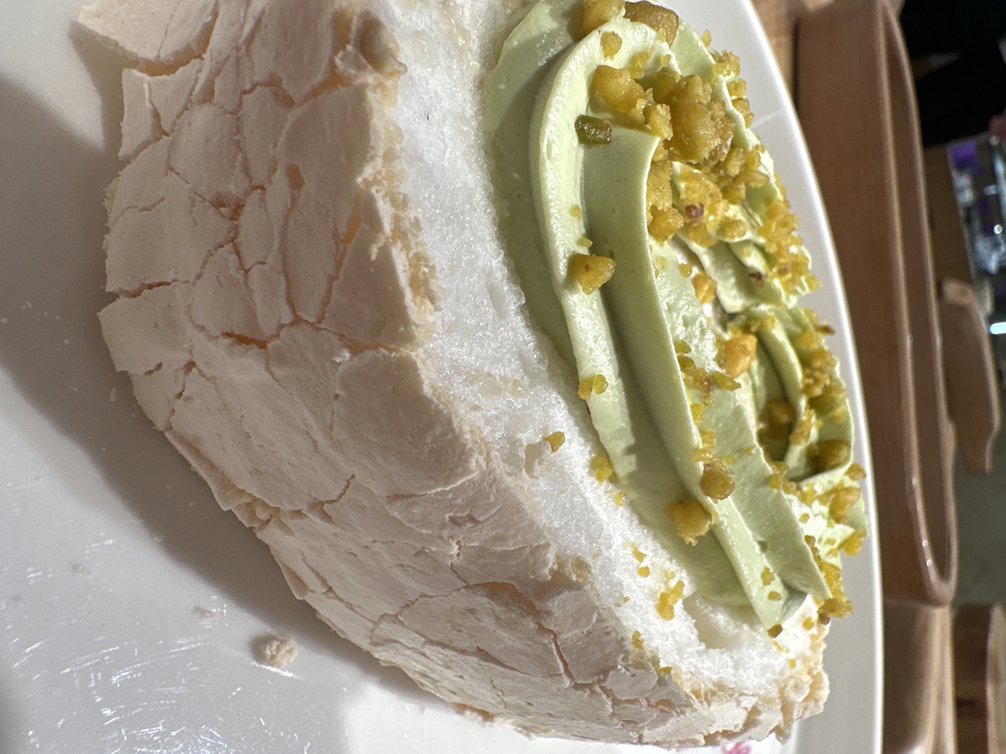 The image shows a close-up of a dessert resembling a Pavlova. The base appears to be a white, meringue-like shell with a cracked texture. On top of the base are swirls of light green cream. The cream is sprinkled with chopped nuts, potentially pistachios. The dessert sits on a white plate. In the background, out of focus, are parts of what appear to be a wooden structure and potentially a glimpse of a window. There is no direct evidence in the image to definitively confirm the location in Rostov-on-Don, Russia, or any specific details about people or interactions.
