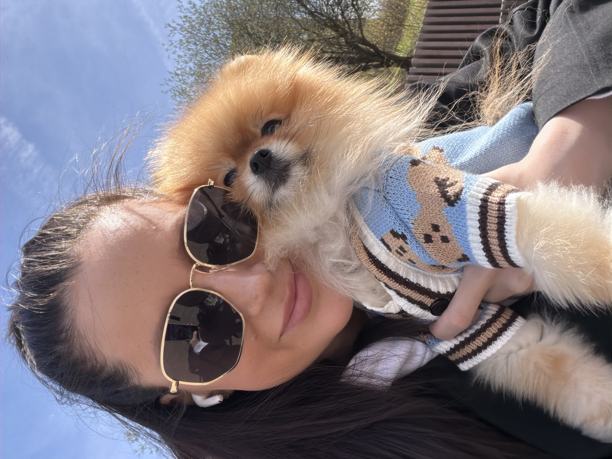 A woman is holding a Pomeranian dog outdoors. The dog is wearing a blue and brown sweater. The woman is wearing sunglasses and has an earbud in her ear. The location is not discernible as being Khimki, Russia from this image alone. The sky is blue with some trees visible in the background.