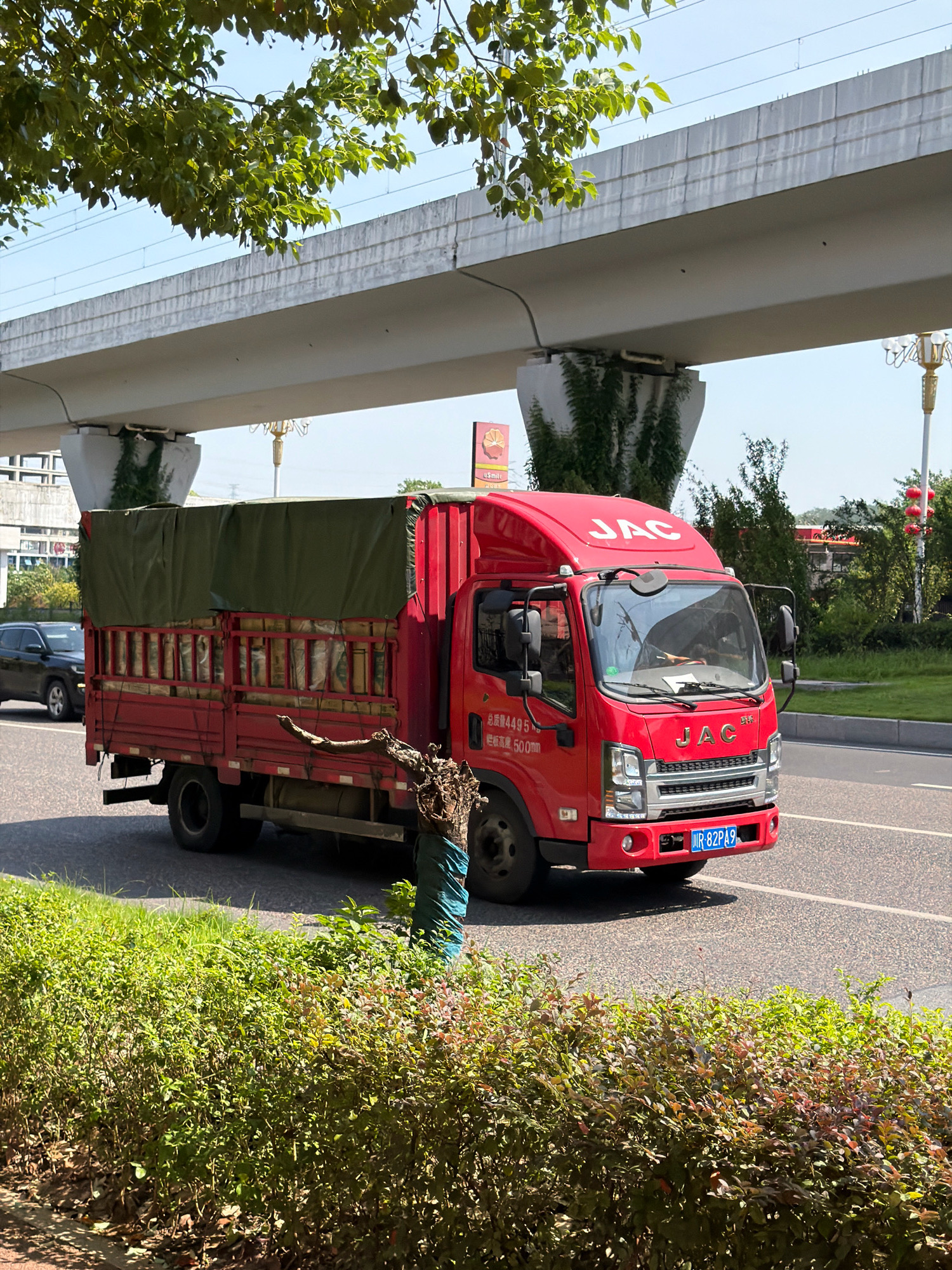 A red JAC truck is driving on a road in Jiangjin, China. The truck is covered with a green tarp, and it appears to be carrying goods. The truck has a license plate that reads "JR 82PA9". In the background, there is an elevated road or bridge, and some buildings and trees. The sky is clear and blue. The truck is traveling on the right side of the road, suggesting this is a country that drives on the right.
