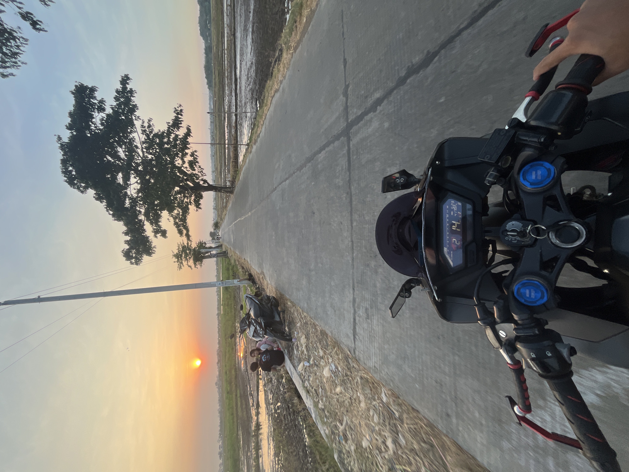 A first-person perspective shows a person riding a motorcycle down a paved road in Pecangaan, Indonesia, at sunset. The sun is low on the horizon, casting a warm orange glow across the sky and reflecting in the water-filled rice paddies on the left side of the road. A few people are sitting on a low wall beside the road, next to two parked motorcycles. A large tree stands on the right side of the road, its silhouette dark against the sky. The motorcycle's dashboard displays the time as 17:14 and the date as 12 14. The road is lined with vegetation and leads into the distance.