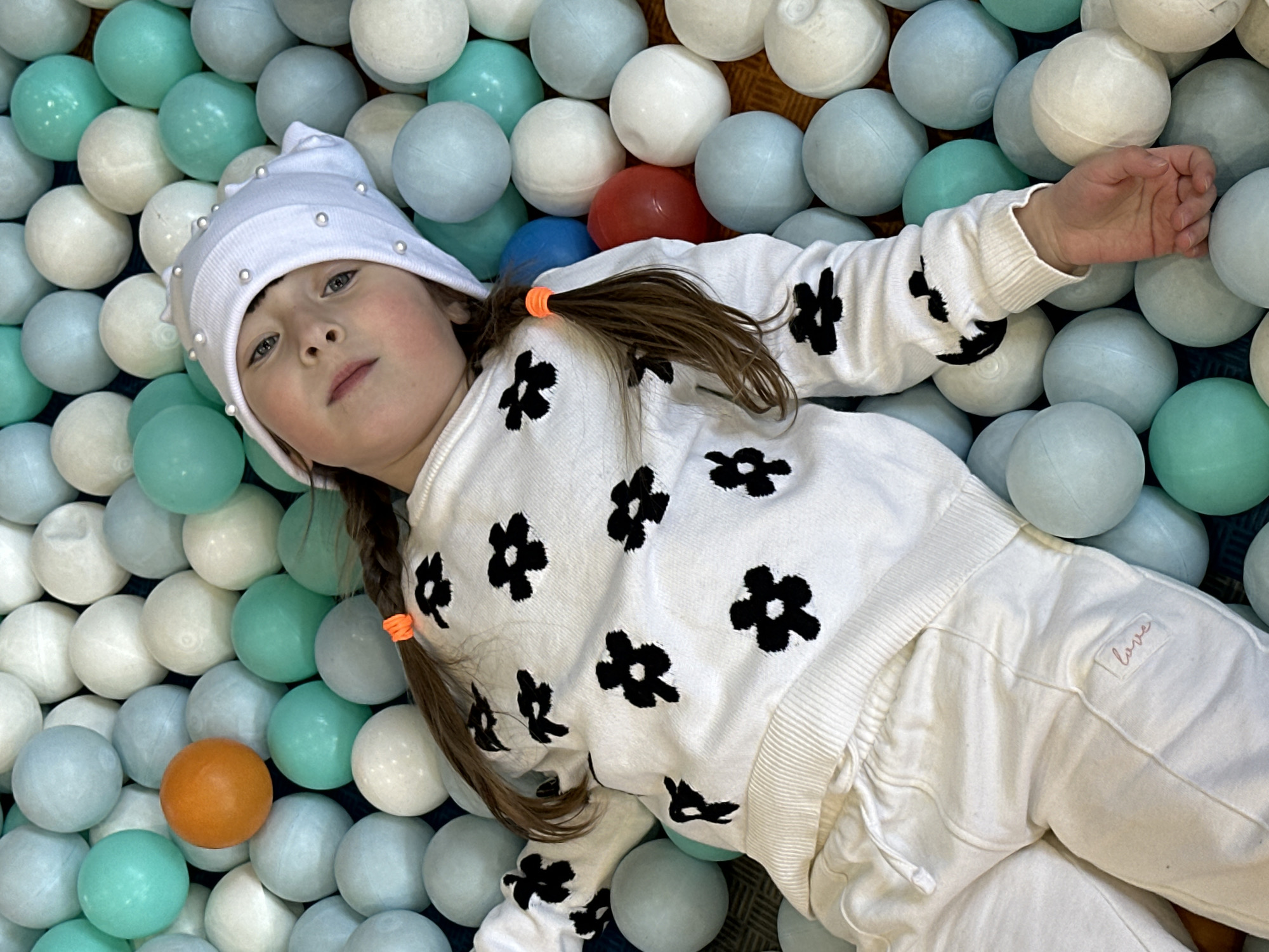 A young child is shown lying on their back in a colorful ball pit in Sochi, Russia. The child, appearing to be a girl, has fair skin, light-colored eyes, and brown hair styled into two pigtails secured with bright orange elastic bands. She is wearing a white knit beanie adorned with small silver or pearl-like beads, a white long-sleeved sweatshirt featuring multiple black floral or star-shaped patterns, and matching white pants.
Her gaze is directed towards the camera with a calm, neutral, or slightly contemplative expression. Her left arm is extended and resting among the balls. The ball pit is filled with numerous plastic balls in a soothing palette of white, light blue, and teal/mint green. A few distinct red, blue, and orange balls are scattered throughout the pit, providing small pops of contrasting color. On the upper right leg of her white pants, a small rectangular patch with the word "love" written in light pink cursive script is visible. The scene captures a moment of rest or play within an indoor entertainment or play area.