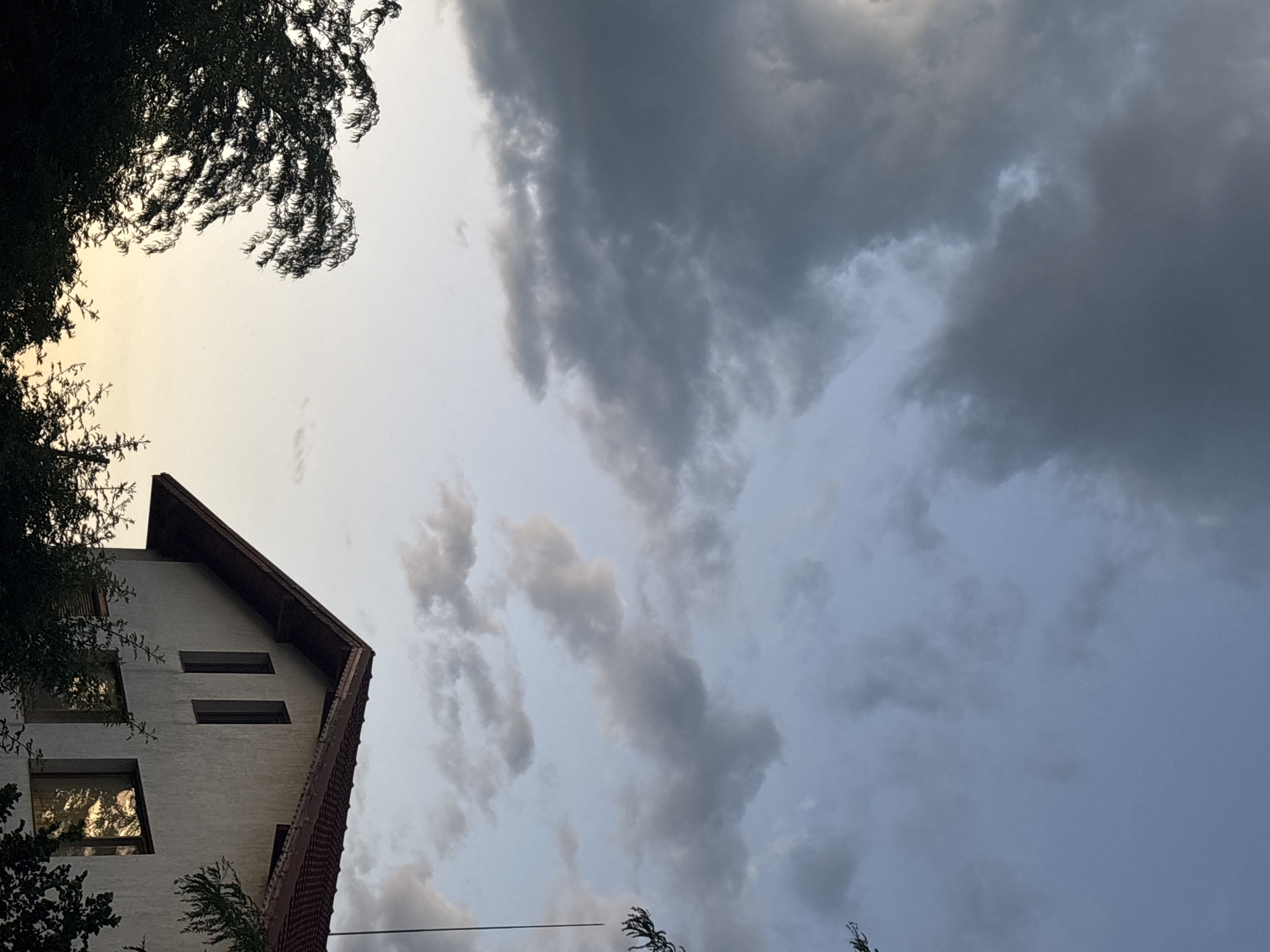 This is a low-angle, vertical shot of a building and the sky. On the left side, the top of a building with a white facade and a dark red tiled roof is visible. The building has several windows, some of which reflect the sky and trees. Trees with dark green foliage are on the far left and bottom left, partially obscuring the building and the sky.
The sky takes up most of the frame and is filled with dramatic clouds. The clouds range in color from dark gray to lighter grays and white, suggesting an overcast or impending storm. The lighting indicates it is either early morning or late afternoon, with a softer, diffused light. The overall mood is somewhat somber due to the cloudy sky, but the reflections in the windows add a touch of brightness.
The scene appears to be in an urban or suburban setting, given the presence of the building. While no people are visible, the building suggests human habitation. The weather is cloudy, possibly threatening rain. The location context is Sofia, Bulgaria. No visible text is present in the image.