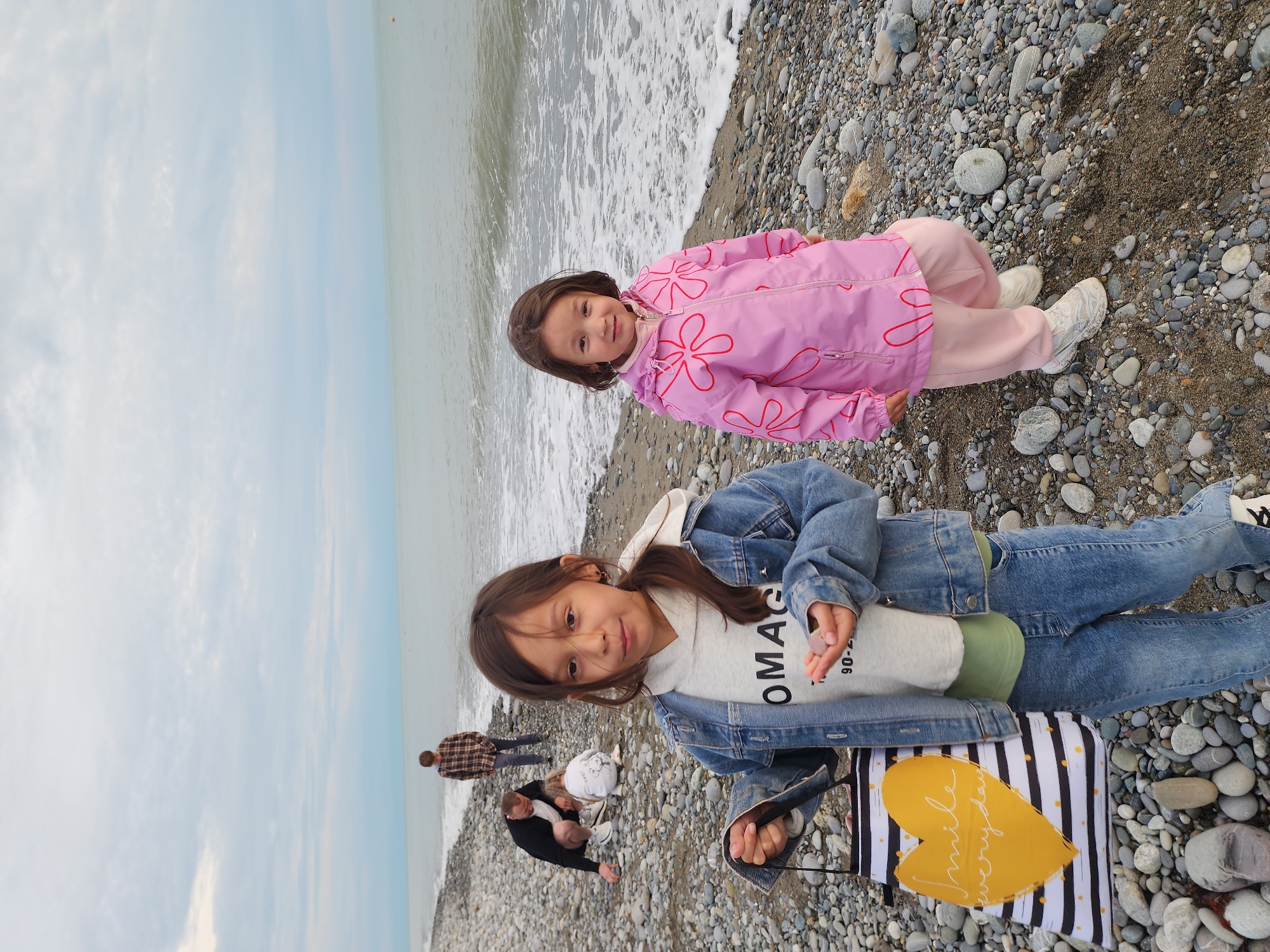 The image depicts a cheerful scene on a pebble beach in Sochi, Russia, under an overcast sky. In the foreground, two young girls are posing by the water's edge.
The girl on the left wears a blue denim jacket over a light-colored hooded sweatshirt with the visible partial text "OMAG" and possibly "90-06" further down, paired with blue jeans. She has long brown hair and is holding a small pebble in her right hand, while also carrying a black and white striped tote bag featuring a large yellow heart with the inscription "smile everyday" on its front.
To her right, a younger girl stands closer to the water, dressed in a bright pink hooded jacket with red floral patterns, light pink pants, and white sneakers. She has shorter brown hair and is smiling at the camera.
The background features the calm, greyish-blue sea with gentle white-foamed waves lapping onto the shore, which is entirely covered in smooth, grey pebbles. Further along the beach, two adults are visible: one standing in a plaid shirt, and another in a dark jacket, appearing to be stooping or kneeling. The sky is largely cloudy, suggesting an overcast daytime. Both girls appear happy and relaxed, enjoying their time on the beach.