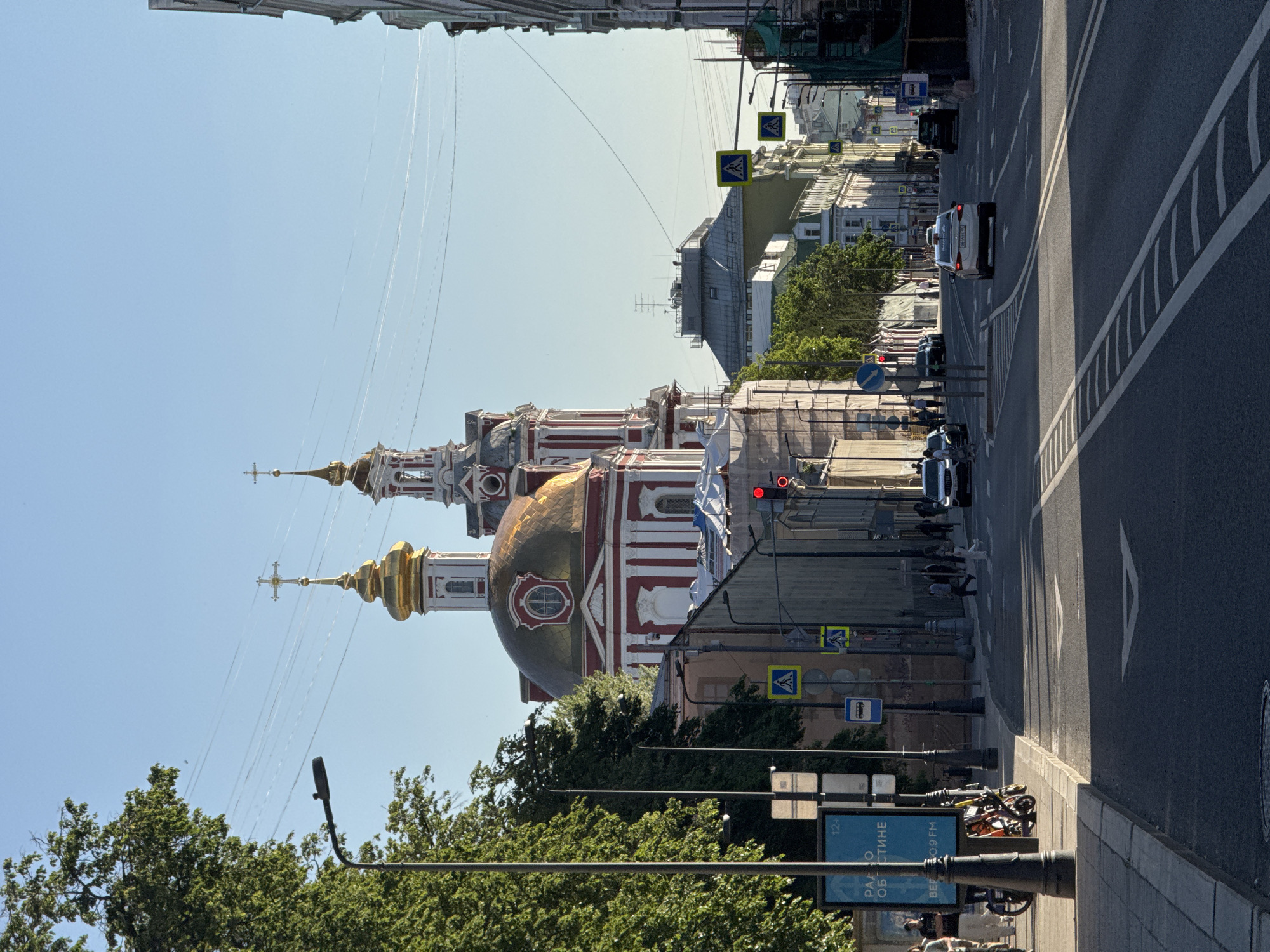 This image captures an urban street scene in Moscow, Russia, featuring the Church of the Great Ascension at Nikitskiye Vorota. The photograph is taken on a clear, sunny day, evidenced by the bright blue sky and distinct long shadows, suggesting either early morning or late afternoon. The most prominent subject is the **Church of the Great Ascension at Nikitskiye Vorota**, located centrally in the middle distance. It is an Orthodox church characterized by its red and white striped facade, white pilasters, and a large golden dome with a drum, topped with an Orthodox cross. To its right, a smaller bell tower with a matching golden dome and cross is visible. The church is well-lit by the sun. The **street**, paved with asphalt, occupies the foreground and extends into the distance. It features clear white lane markings, including solid and dashed lines, and a white arrow indicating a straight-ahead direction in the right lane. A central reservation or median is marked by parallel dashed lines. Several **vehicles** are present. A white sedan (possibly a Hyundai) is moving away from the viewer in the right lane, centered in the mid-ground. Further down the street on the right, a white SUV is partially visible, appearing to be stopped or moving slowly. Other cars are visible in the background along both sides of the street. Multiple **pedestrians** are observed. One individual in a light top and dark trousers is crossing the street from right to left in the middle distance. Others are visible walking on the sidewalks on both sides of the street. On the far left sidewalk, a person is walking near a large blue **billboard** displaying Russian text "РАДИО ОБЛАСТИ" (Radio of the Region) and "BEP 9.0.9 FM", along with a "12+" age restriction sign. Various **street furniture** and **signs** are interspersed throughout the scene. Black lampposts line both sides of the street. A red traffic light is visible on the right side. Yellow diamond-shaped pedestrian crossing signs with black pedestrian icons are present on both sides. A blue square sign with a white bus icon (indicating a bus stop) is seen on the left sidewalk. Several bicycles or electric scooters are parked on the left sidewalk near the billboard. Buildings of varying architectural styles line both sides of the street. Some buildings, particularly on the right, appear to be undergoing renovation or construction, indicated by scaffolding and protective netting. Numerous thin