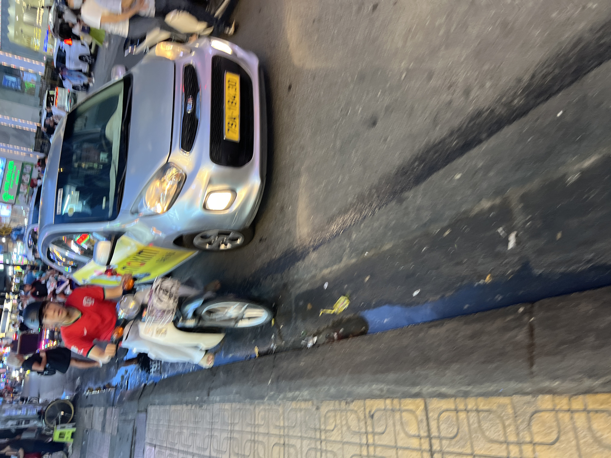 The image depicts a street scene in Nha Trang, Vietnam. A young man wearing a red shirt and white pants is riding a white scooter. He is wearing a black helmet and appears to be looking at the camera. Beside him, a silver car is partially visible. The car has a yellow license plate that reads "79A-134.30". The street is wet and appears to be a busy area with other vehicles and pedestrians in the background. There is a sidewalk on the left side of the image with a patterned pavement. The overall lighting suggests it is daytime or early evening.