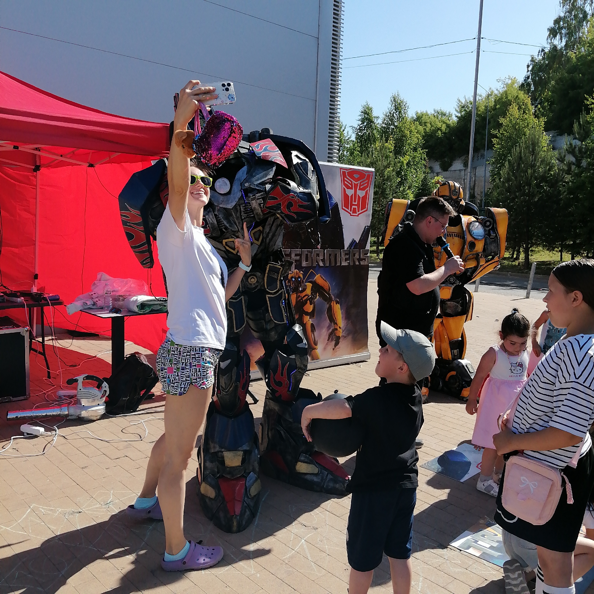 The image depicts an outdoor event taking place in Yudino, Russia, under bright daylight with a clear blue sky. The scene is set on light-colored paved ground, where several individuals interact with two large robot costumes resembling Transformers characters. In the left-center, a fair-skinned young adult female, wearing a white t-shirt, brightly patterned shorts, purple Crocs with light blue socks, and neon yellow sunglasses, holds a white smartphone aloft with her left hand, appearing to take a selfie or photograph. A small brown plush toy and a pink sequined pouch hang from her wrist. She smiles broadly, revealing teeth. Her right hand shows a light blue wristband. She stands next to a tall, dark-colored robot costume resembling Optimus Prime, featuring blue, red, and black components with flame motifs. To the right of Optimus Prime, a vertical banner displays the "TRANSFORMERS" logo and an image of Bumblebee, topped by the Autobot symbol. Behind this banner and further right, another large robot costume, predominantly yellow and resembling Bumblebee, stands. In front of the Bumblebee costume, an adult male with glasses, dressed in a black polo shirt, holds a black microphone in his right hand, seemingly addressing an audience. In the foreground, a young boy wearing a dark baseball cap, black t-shirt, and dark shorts looks upwards and to the right, holding a black spherical object. Behind him, a younger girl in a pink "Hello Kitty" dress and white sandals looks downwards. To the far right, an older girl, wearing a striped t-shirt, black skirt, striped white socks, and grey shoes, carries a small pink shoulder bag with a bow. She is looking to her left, seemingly engaged in conversation. In the left background, a red pop-up canopy tent provides shade. Underneath, two black tables are visible. The left table holds various wires and electronic equipment, while the right table has some wrapped items and bottles. A white piece of equipment, possibly a fan, is on the ground near the tables, surrounded by numerous electrical cables. In the background, behind the tent and costumes, a large, light-grey building with vertical ridges is visible. Further back and to the right, a line of green trees and bushes, along with a utility pole and overhead wires, are present against the clear sky. Faint white chalk drawings are visible on the paved ground in various spots. Distinct shadows are cast on the ground, indicating a sunny