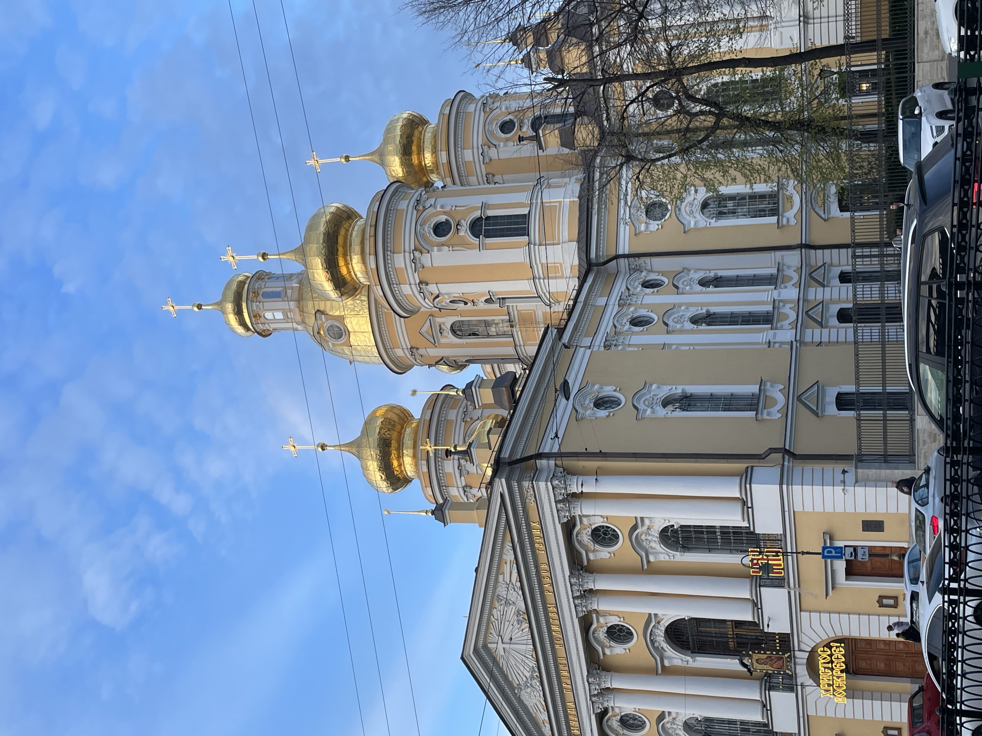 The image shows the Naval Cathedral of Saint Nicholas in Saint Petersburg, Russia. The view is from below, looking up at the cathedral's facade and golden domes against a blue sky with some clouds. Several power lines stretch across the upper portion of the image. The building's architecture features pale yellow walls, white columns, decorative windows, and golden domes topped with Orthodox crosses. Some bare trees are visible on the right side of the frame, along with parked cars and a wrought-iron fence. A sign in Cyrillic script, possibly related to the cathedral, is also visible near the entrance.