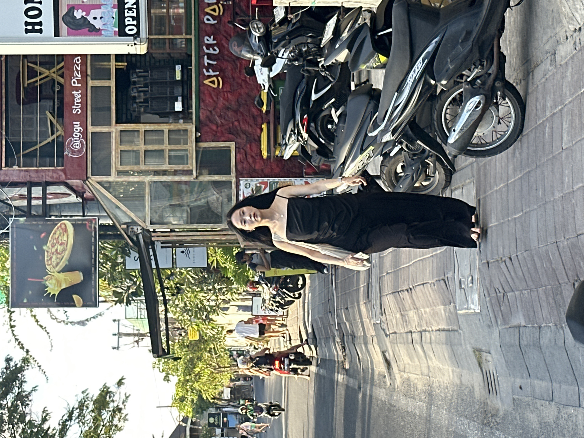 A young woman in a long black dress stands on a sidewalk next to parked motorcycles. She is looking up and to her right, with her left arm outstretched. The scene is a street in North Kuta, Indonesia, likely during the day as indicated by the bright sunlight and shadows. The sidewalk is paved with stone and there are several motorcycles parked along it. In the background, there are buildings with signs advertising "HOM," "OPEN," and "AFTER PARTY." One building has a sign that says "GgGgu Street Pizza" with an image of a pizza. Further down the street, more people can be seen walking and riding motorcycles. The overall atmosphere appears to be casual and lively, typical of a busy street in Southeast Asia.