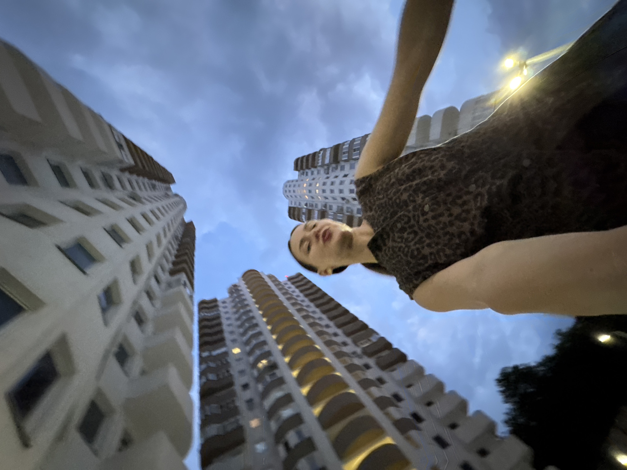 A young woman with dark hair pulled back in a ponytail is pictured from a low-angle perspective, looking upwards. She is wearing a short-sleeved, leopard-print dress. Her right arm is extended towards the camera, and she appears to be taking a selfie or filming herself.
Behind her, two tall, modern apartment buildings dominate the frame. The building on the left is light-colored with balconies featuring yellow accents, and the one on the right is also light-colored with a more curved architectural style, its windows lit up. The sky above is overcast with dark, dramatic clouds, suggesting twilight or a storm is approaching.
The scene takes place outdoors, possibly in a city park or residential area, with glimpses of dark trees and streetlights in the lower portion of the image. The overall impression is one of urban living with a focus on the architecture of the apartment complexes. The image is a wide shot with the woman as the central subject, but the imposing buildings and the sky create a sense of scale and drama. The slight blurriness of the image suggests it was taken at dusk or in low light conditions.