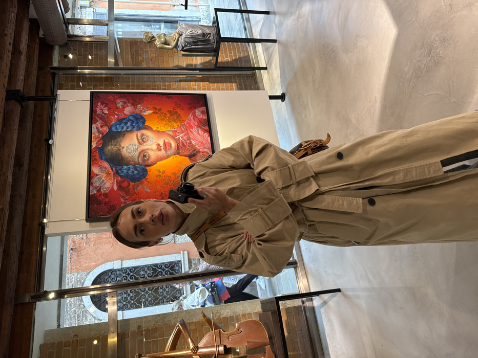A mid-shot depicts a woman standing in an indoor art gallery or exhibition space, looking directly towards the viewer with her mouth slightly ajar. She is wearing a long beige trench coat and holds a small black camera in her left hand. A brown leather shoulder bag is visible on her right hip.
The interior features a polished light gray floor and a ceiling with exposed dark wooden beams. Track lighting illuminates the space. Behind the woman, a large, vividly colored rectangular painting is displayed on a white freestanding partition. The artwork portrays a female face with intricate details, blue braided hair styled into buns, and floral patterns against a vibrant red and orange background. To the left of the painting, a stylized metallic sculpture resembling a cello stands on a black pedestal. To the right, a bronze-toned sculpture of a draped, seated female figure rests on another black pedestal.
Large glass windows frame the central artwork, providing views of the exterior. Through the left window, a narrow Venetian calle (alleyway) is visible, characterized by old reddish-orange brick buildings with arched windows and decorative ironwork. A blurred pedestrian can be seen walking in the alley. The right window reveals additional exterior stone paving and building facades, confirming the location in Venice, Italy.